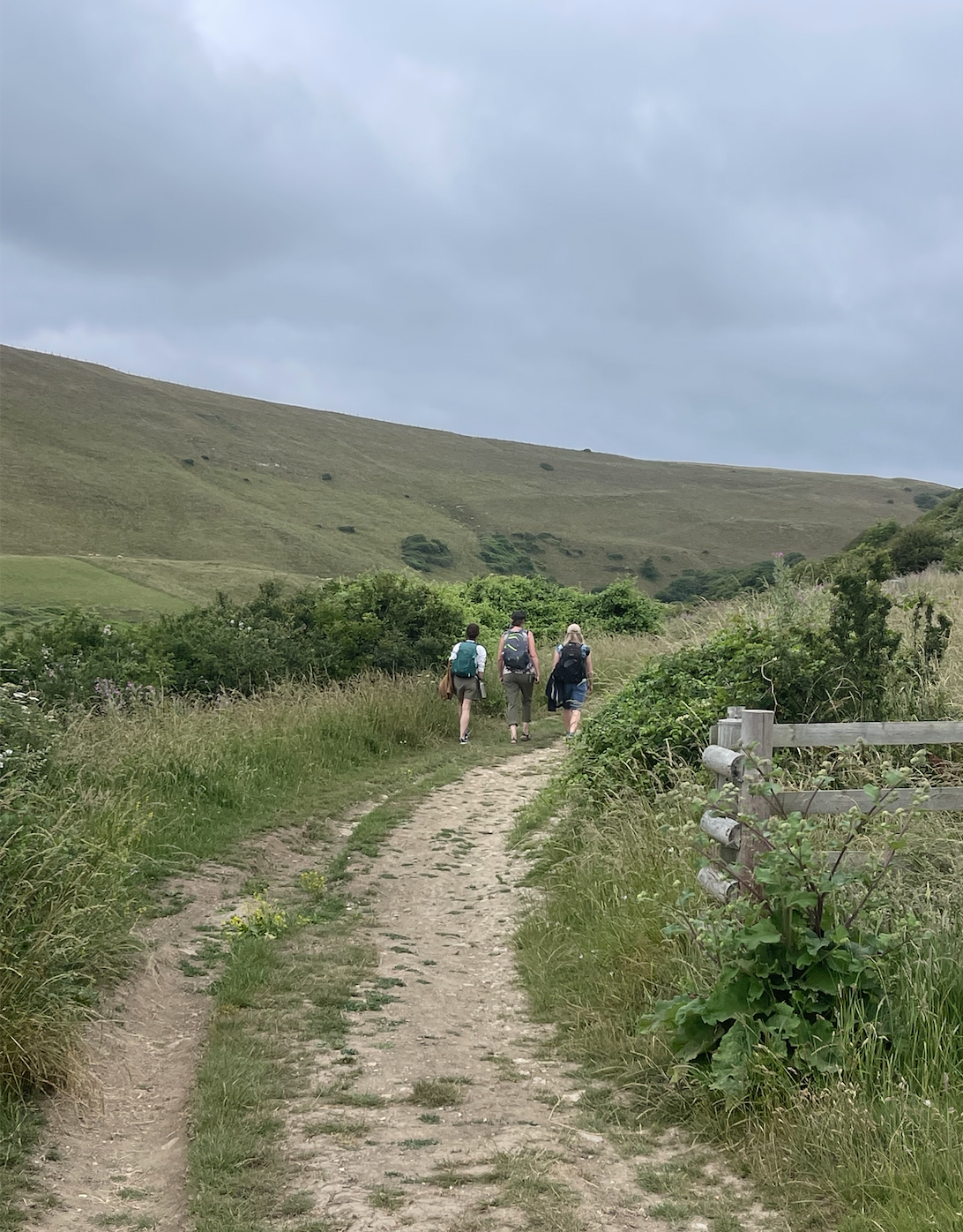 Three people hiking on a dirt trail through a grassy landscape with hills in the background and cloudy sky above.