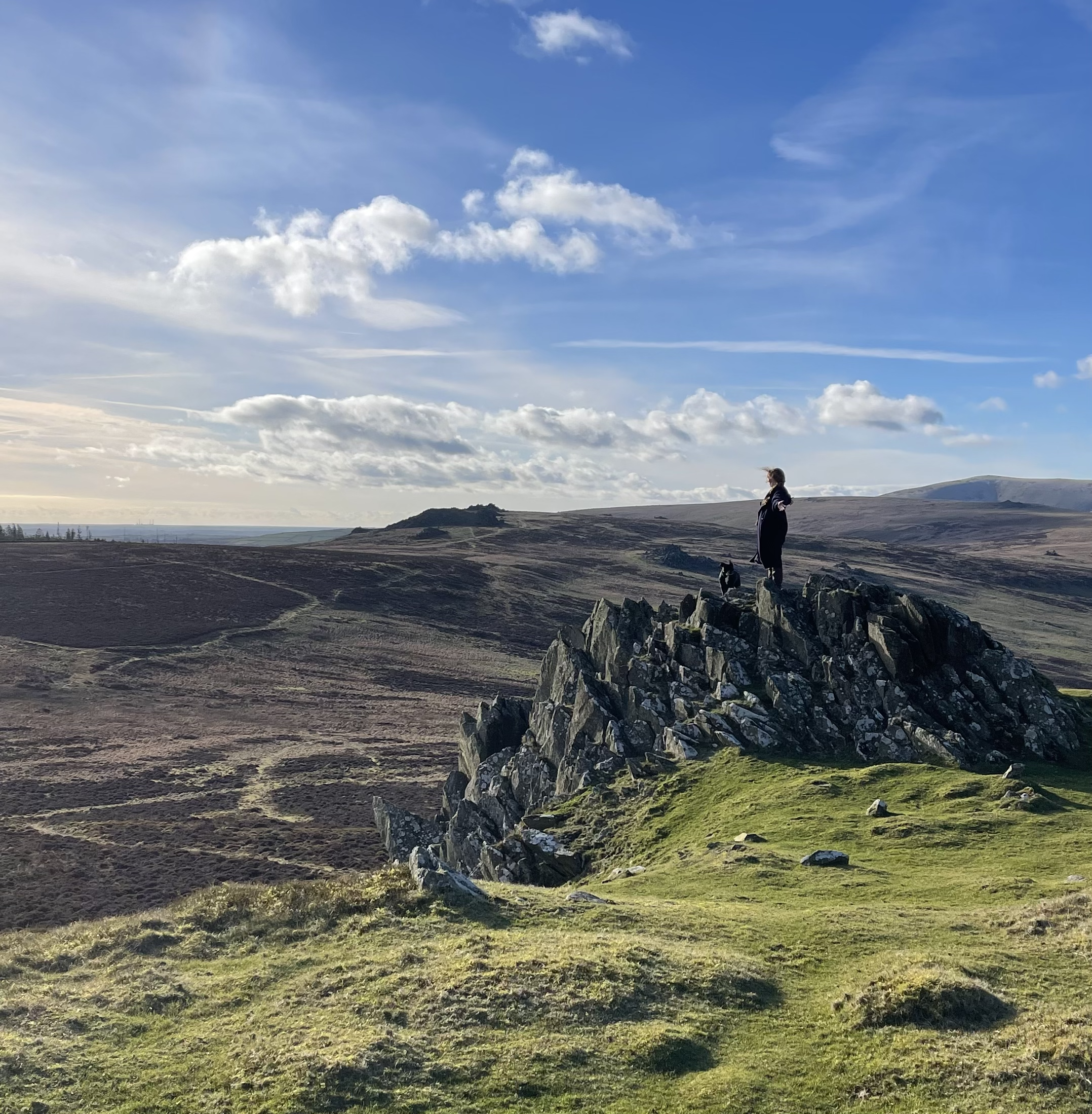 Person and dog standing on a rocky hilltop overlooking a vast landscape of grass and moorland under a cloudy blue sky. Wales