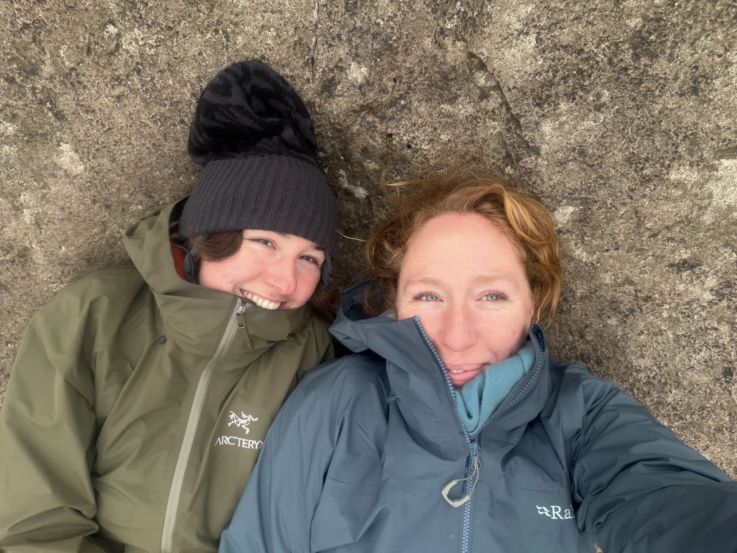 Two women lying on the ground on a rocky surface, smiling and taking a selfie. One woman has red hair and is wearing a blue jacket, the other woman has dark hair, is wearing a black beanie and an olive green jacket.