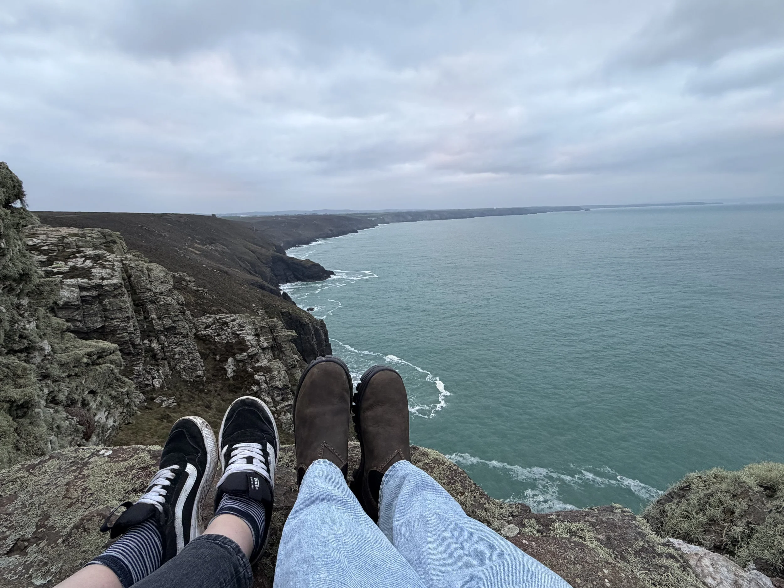 Two pairs of feet, one in sneakers and one in boots, resting on a rocky ledge overlooking an ocean and cliff coastline on a cloudy day.