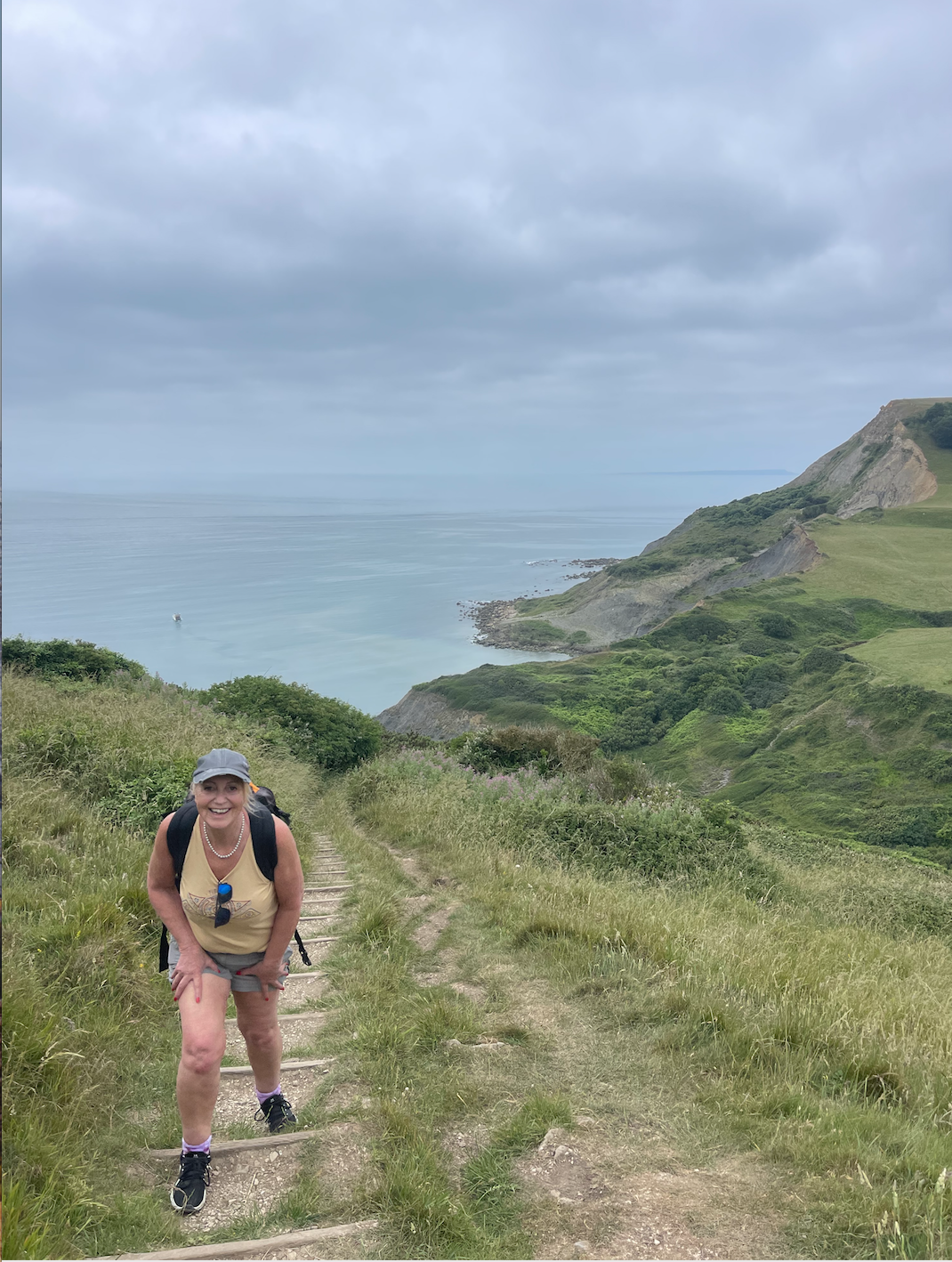 Smiling woman hiking up a grassy trail along a coastal hillside with the ocean and cliffs in the background during overcast weather.