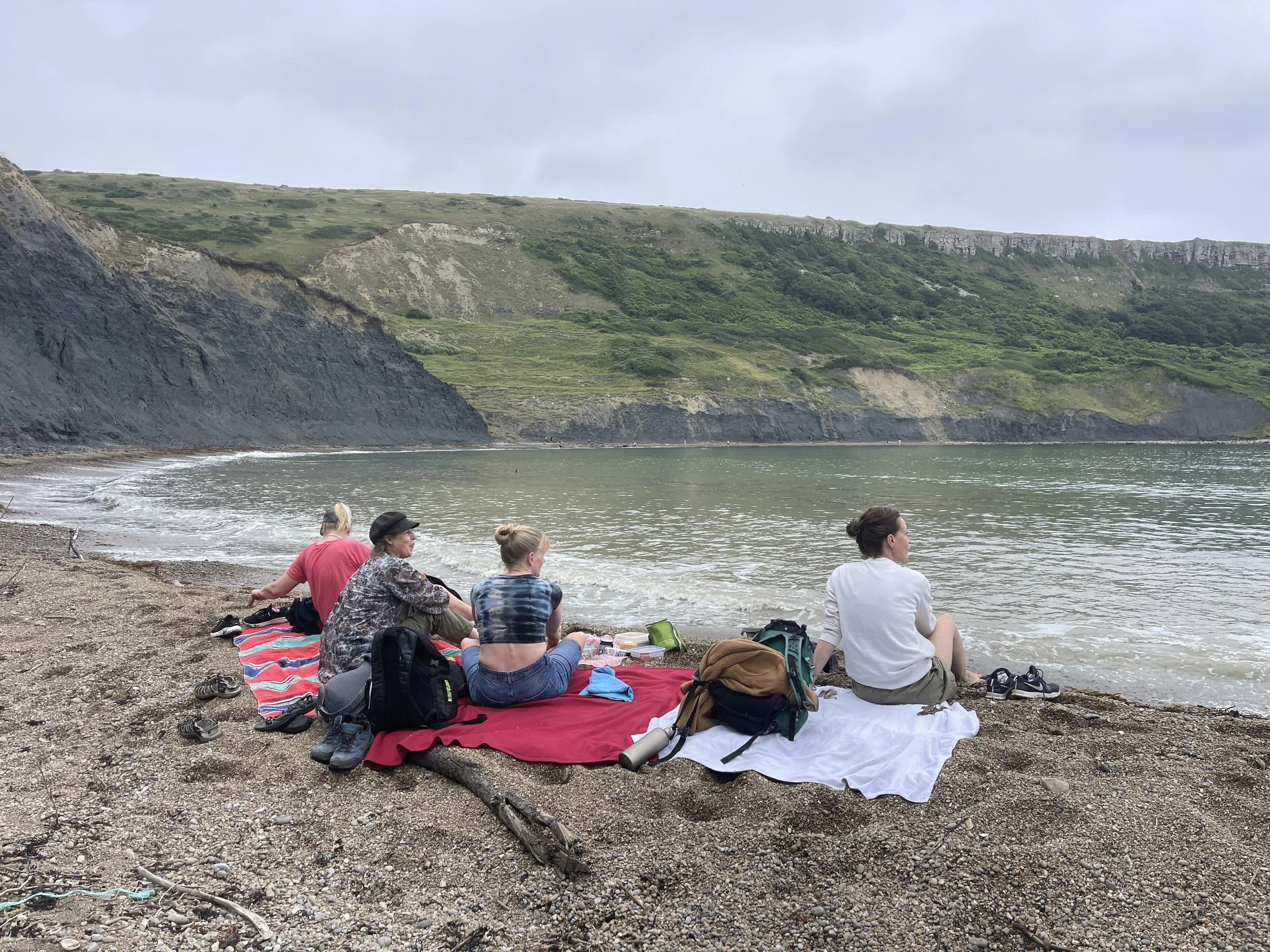 People sitting on towels and blankets on a rocky beach, facing the ocean, with green hills and cliffs in the background.