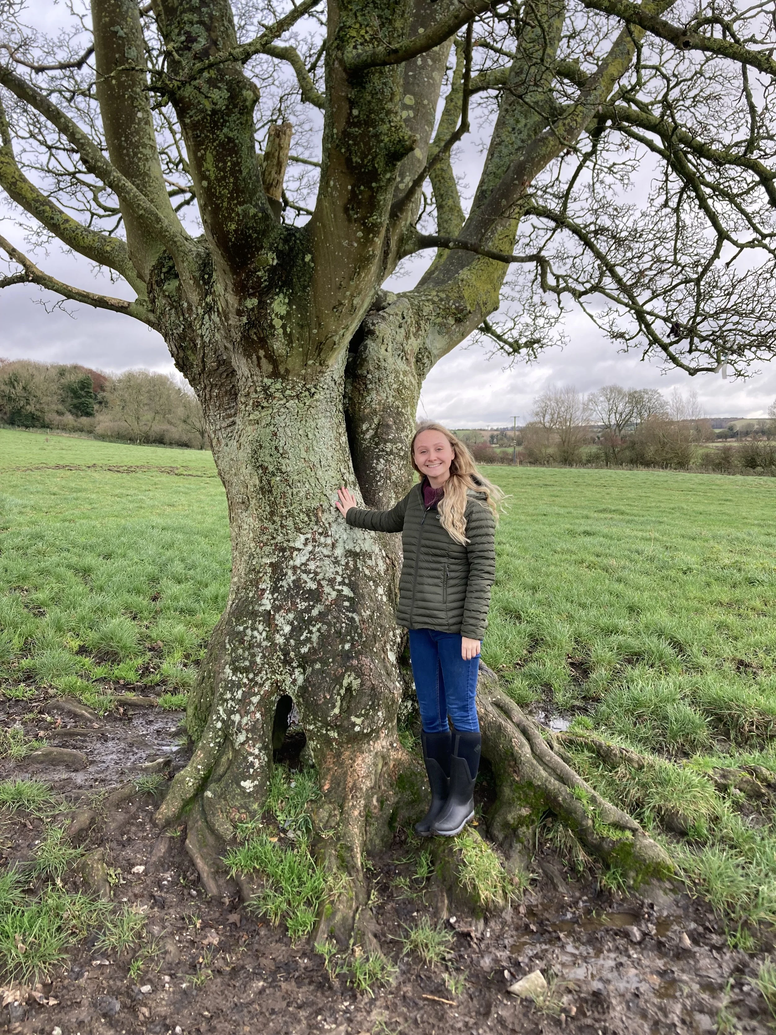 A girl with long blonde hair, wearing a black jacket, blue jeans, and rubber boots, stands next to a large, leafless tree in a grassy field. She is smiling and touching the tree trunk.