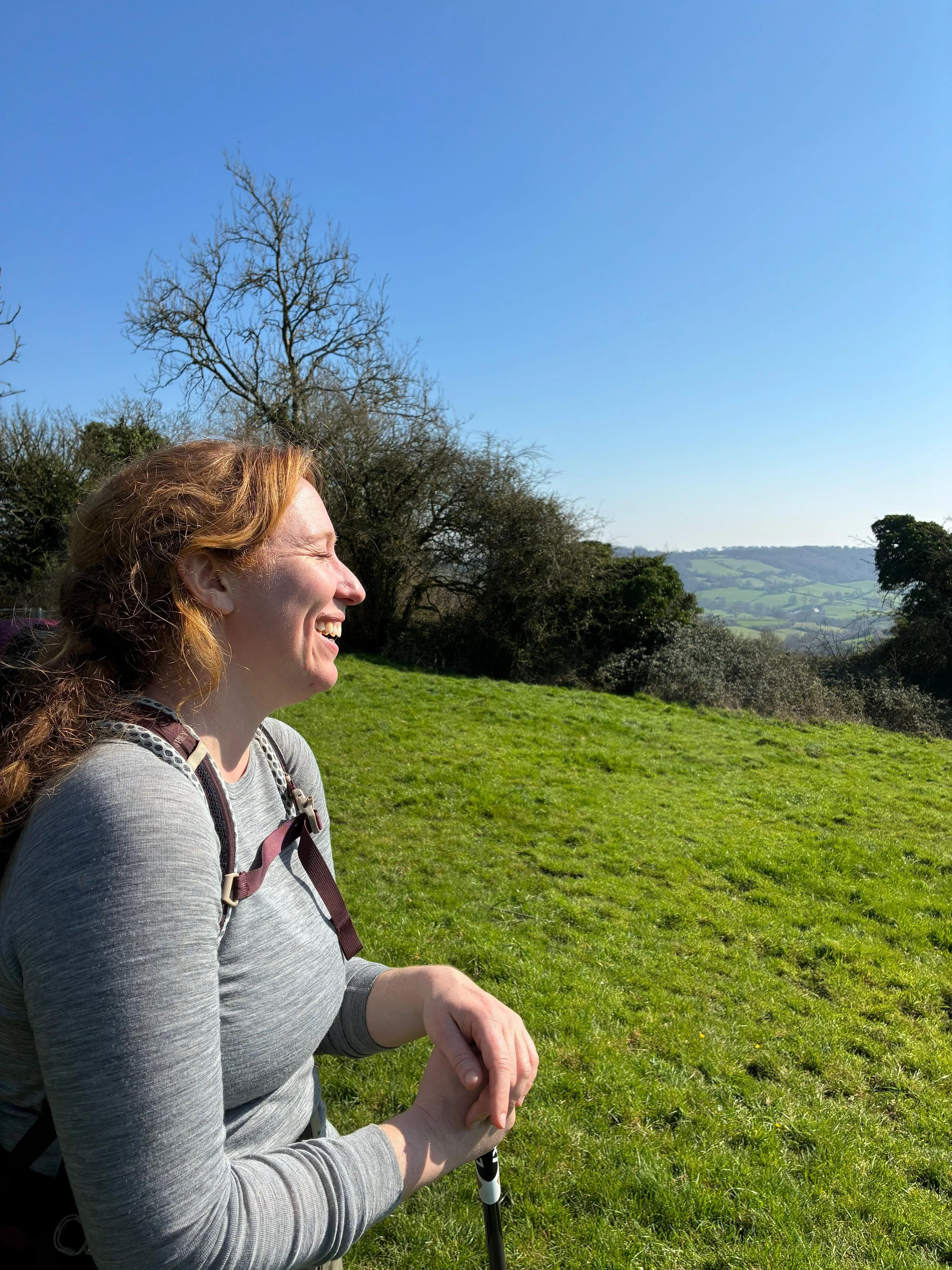 A woman with red hair smiling outdoors on a sunny day, wearing a gray long sleeve shirt and backpack, standing on a grassy hill with trees and rolling hills in the background.