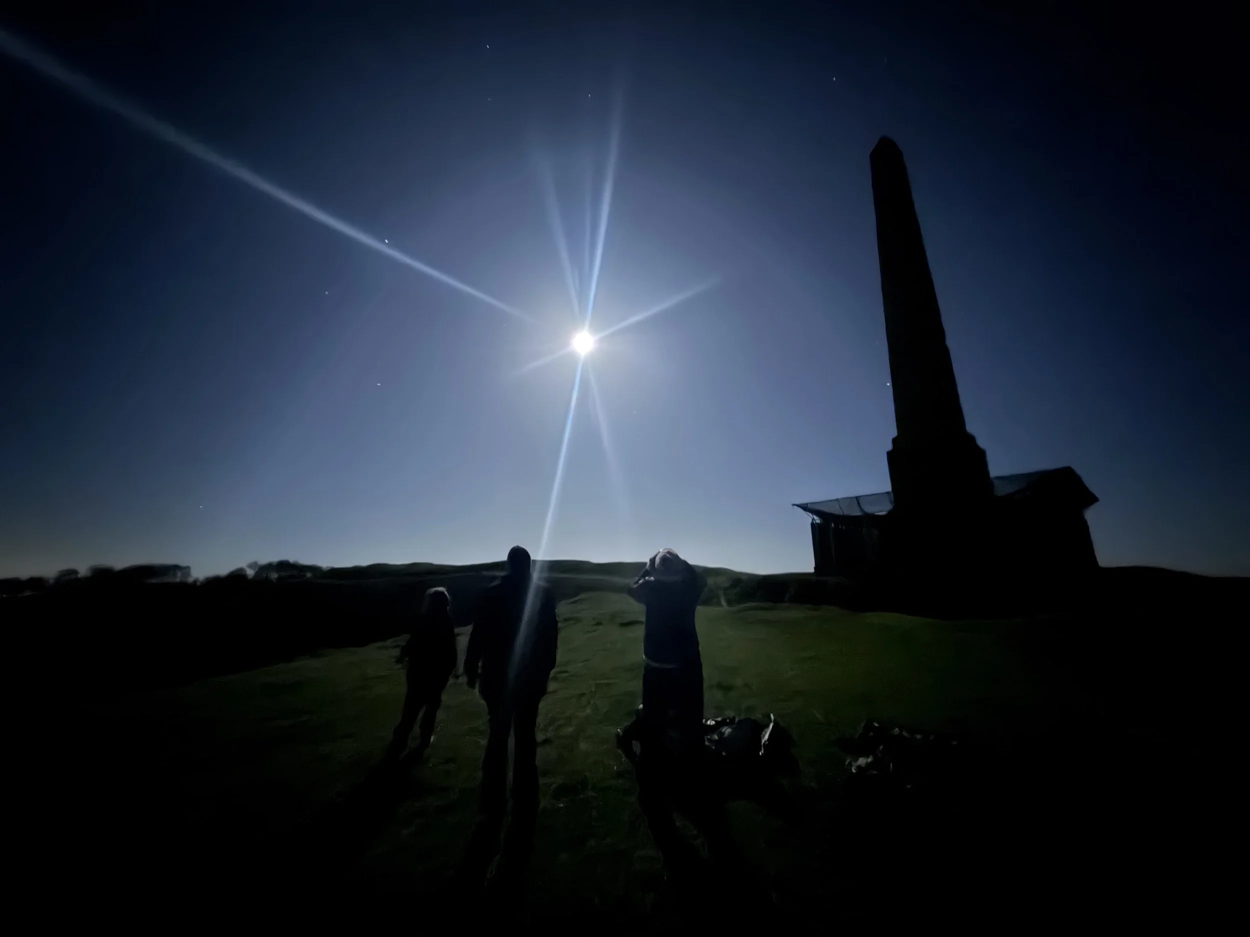 Nighttime scene with three silhouetted people standing on grass, looking up at the moon in the clear sky, with a tall monument or obelisk nearby. Guided full moon night walk, Cherhill White Horse