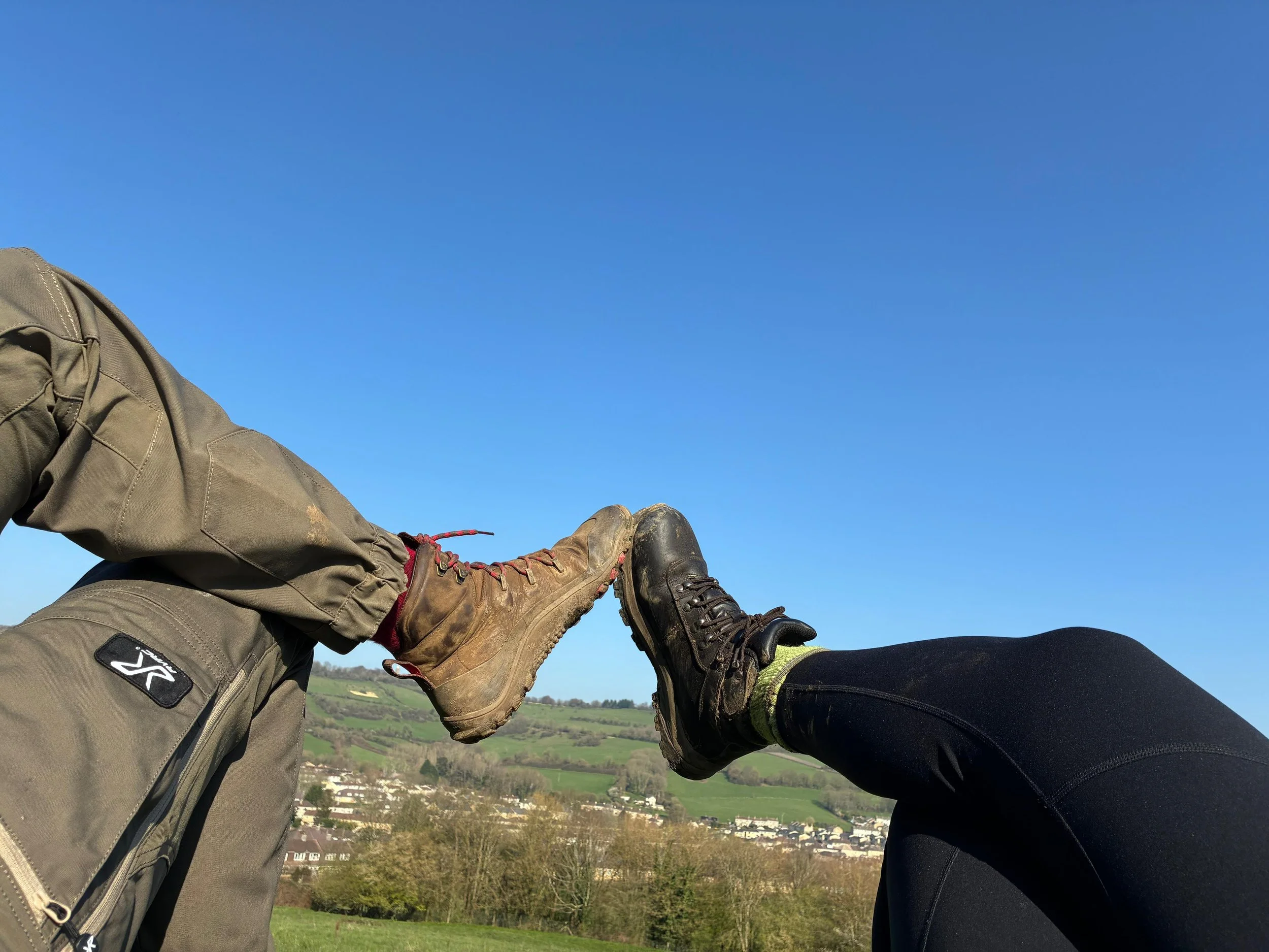 Two people with hiking boots touching feet, outdoors in a green landscape under a blue sky.
