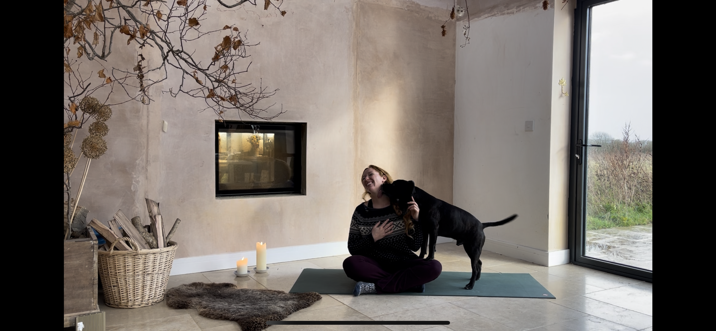 A woman sitting cross-legged on a yoga mat next to a black dog inside a room with beige walls, a glass door showing an outdoor landscape, lit candles, a basket of firewood, a fur rug, and dried branches hanging from the ceiling. Yoga classes, Calne, 