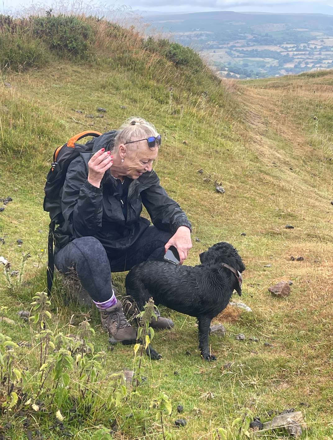 A woman with gray hair, wearing a black jacket, purple socks, and hiking boots, squats on a grassy hillside. She is laughing and holding a smartphone in her right hand. A wet black dog stands beside her, looking up at her with its tongue out. The bac