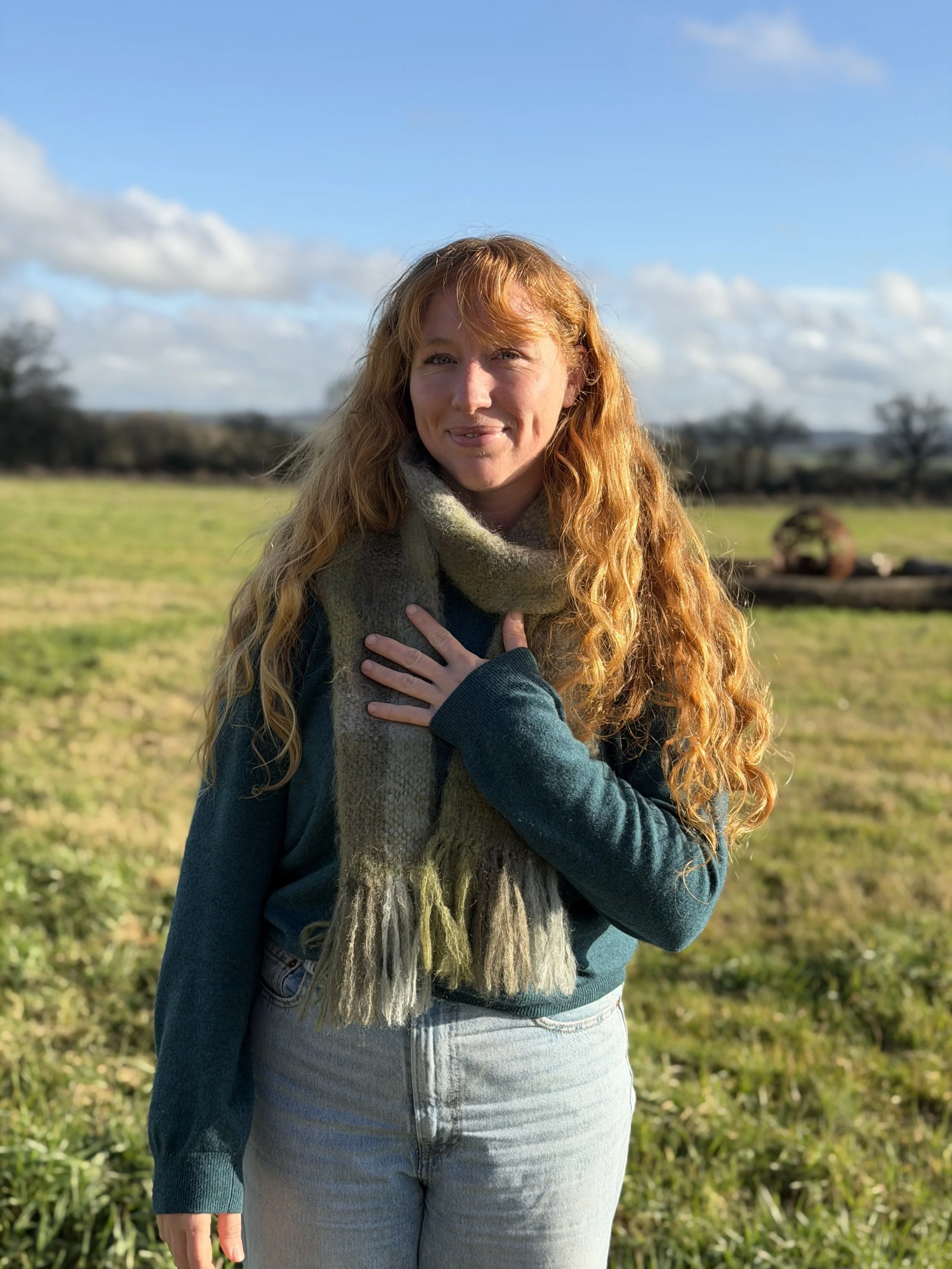 A woman with long red hair standing in a grassy field under a partly cloudy blue sky, wearing a dark sweater, light jeans, and a multicolored scarf, with her hand on her chest.