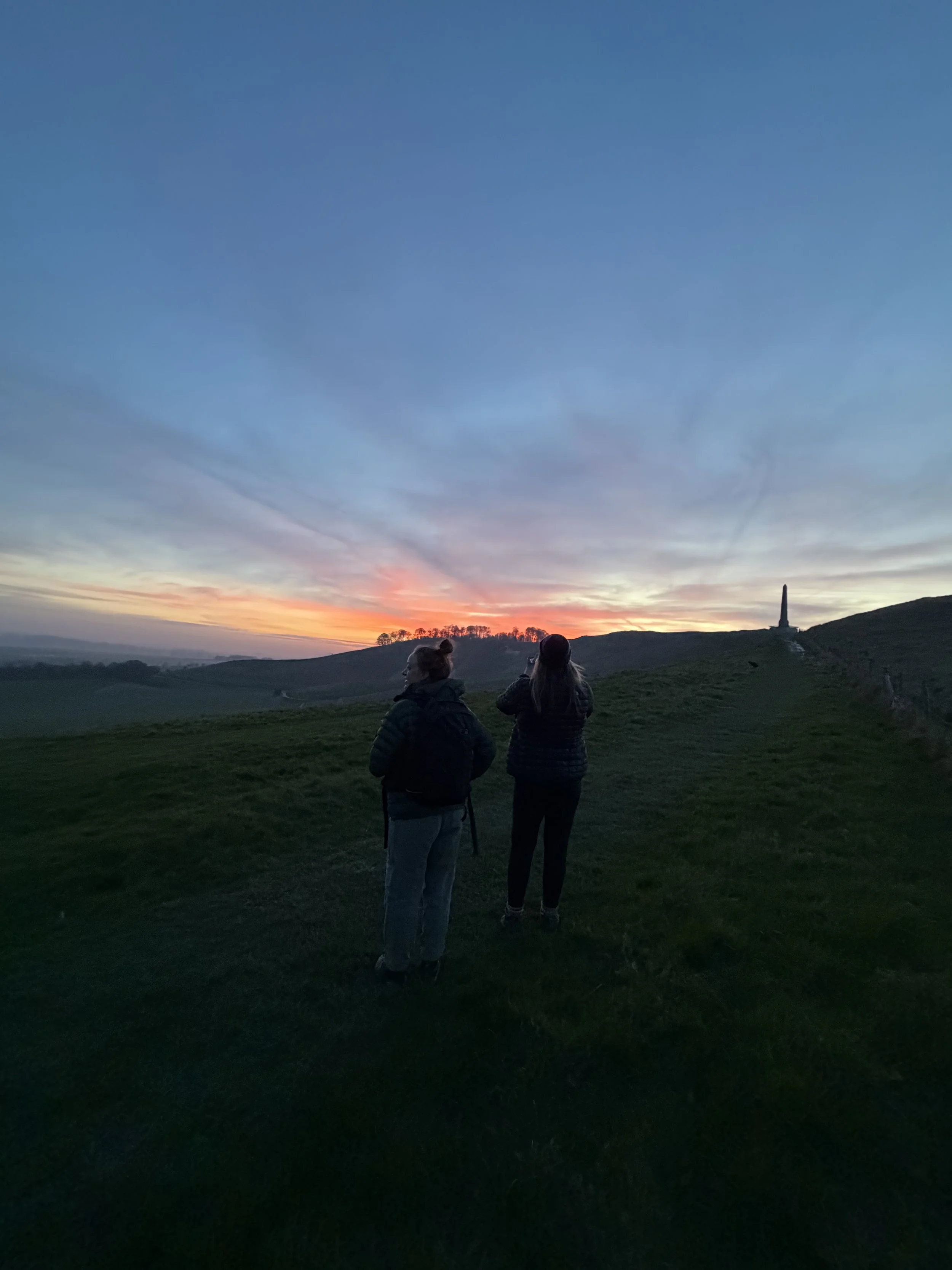 Two women stand on a grassy hill during sunset, with one taking a photo of the scenery featuring a monument on a hillside in the distance.
