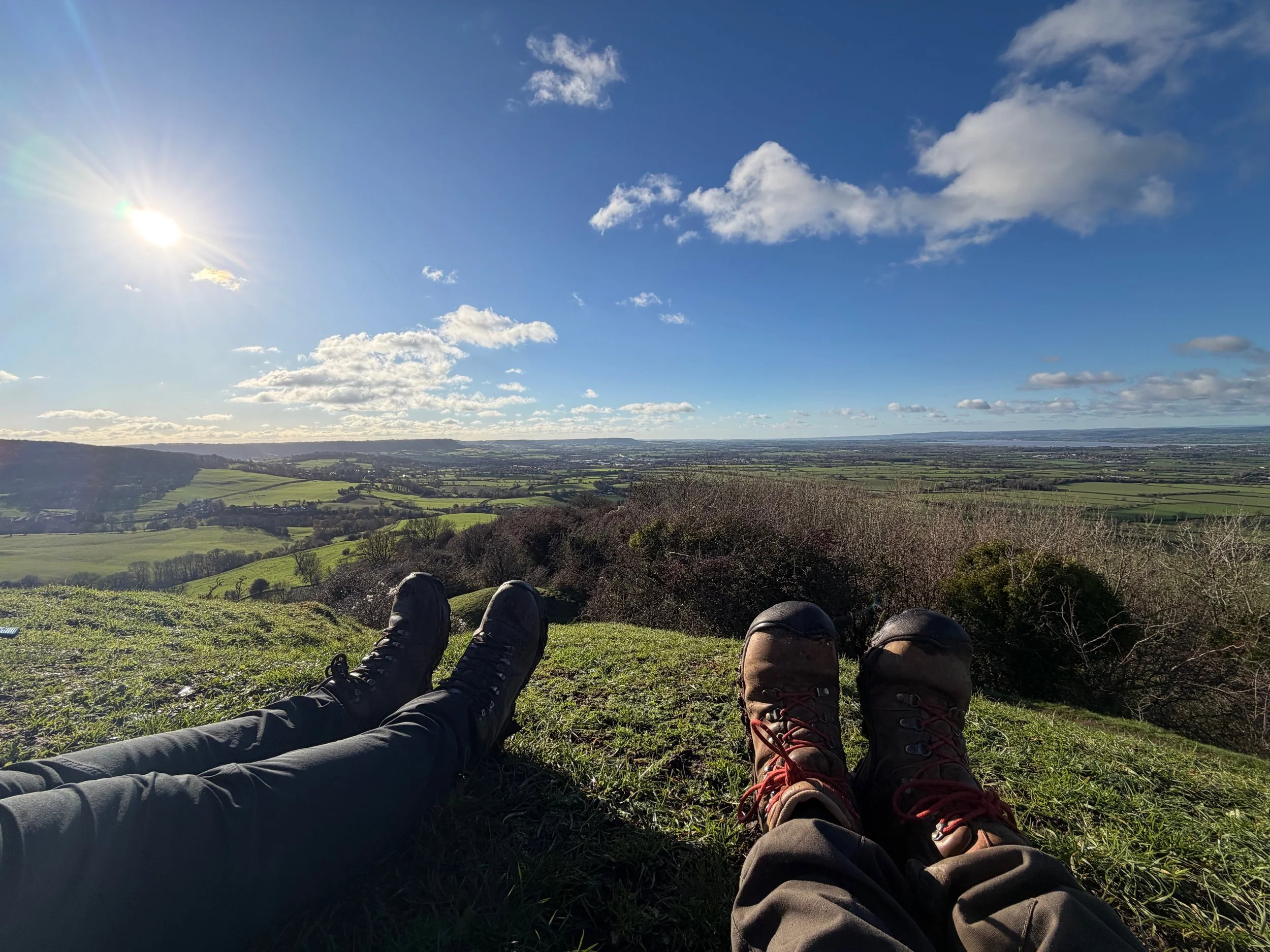 Two people sitting on grass, wearing hiking boots, overlooking a scenic landscape of rolling hills, fields, and trees under a bright blue sky with scattered clouds and the sun shining. Walking the Cotswold Way.