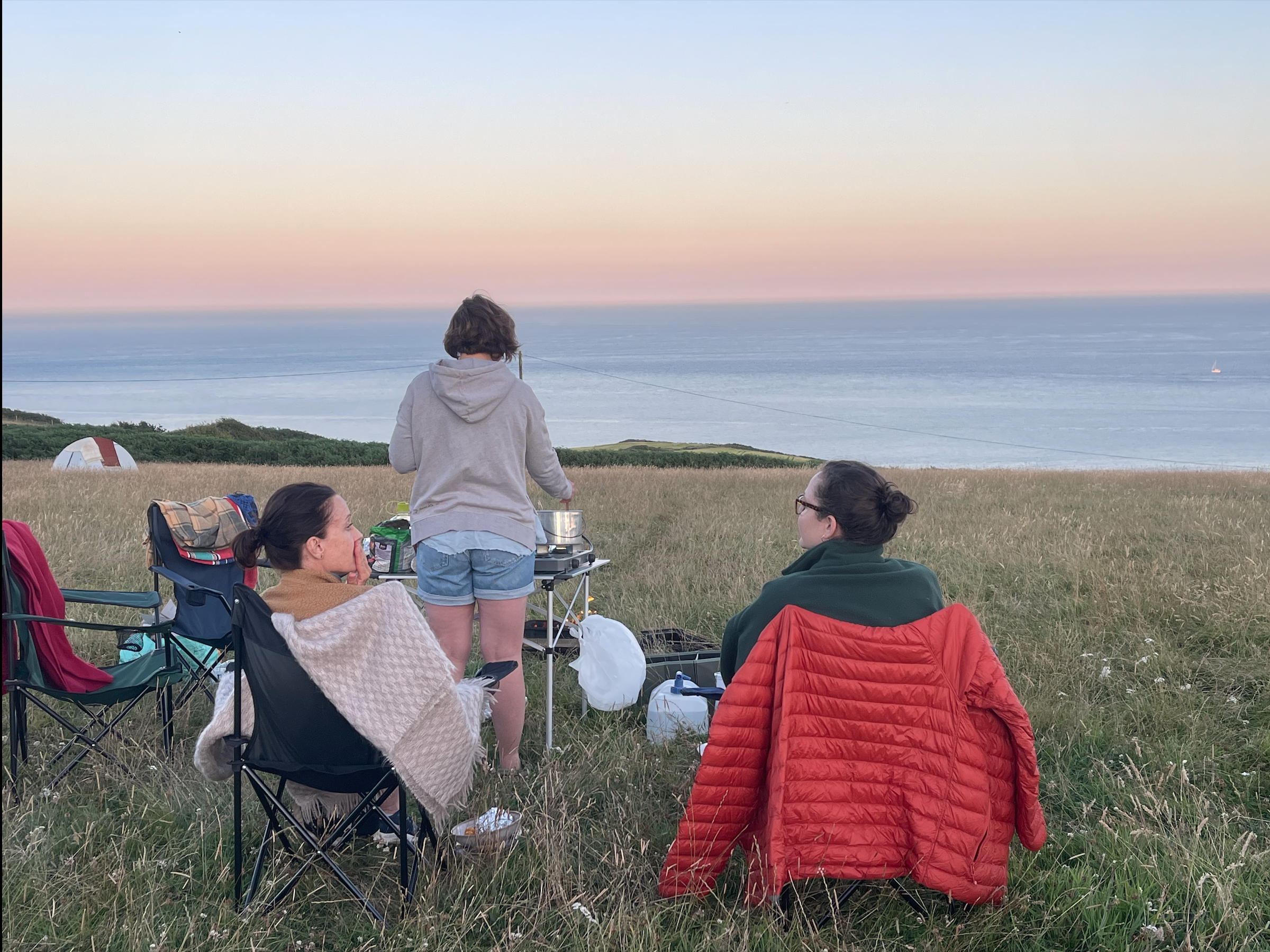 Three women are enjoying an outdoor picnic on a grassy hill overlooking the ocean at sunset, with camping gear and a small table in the scene.