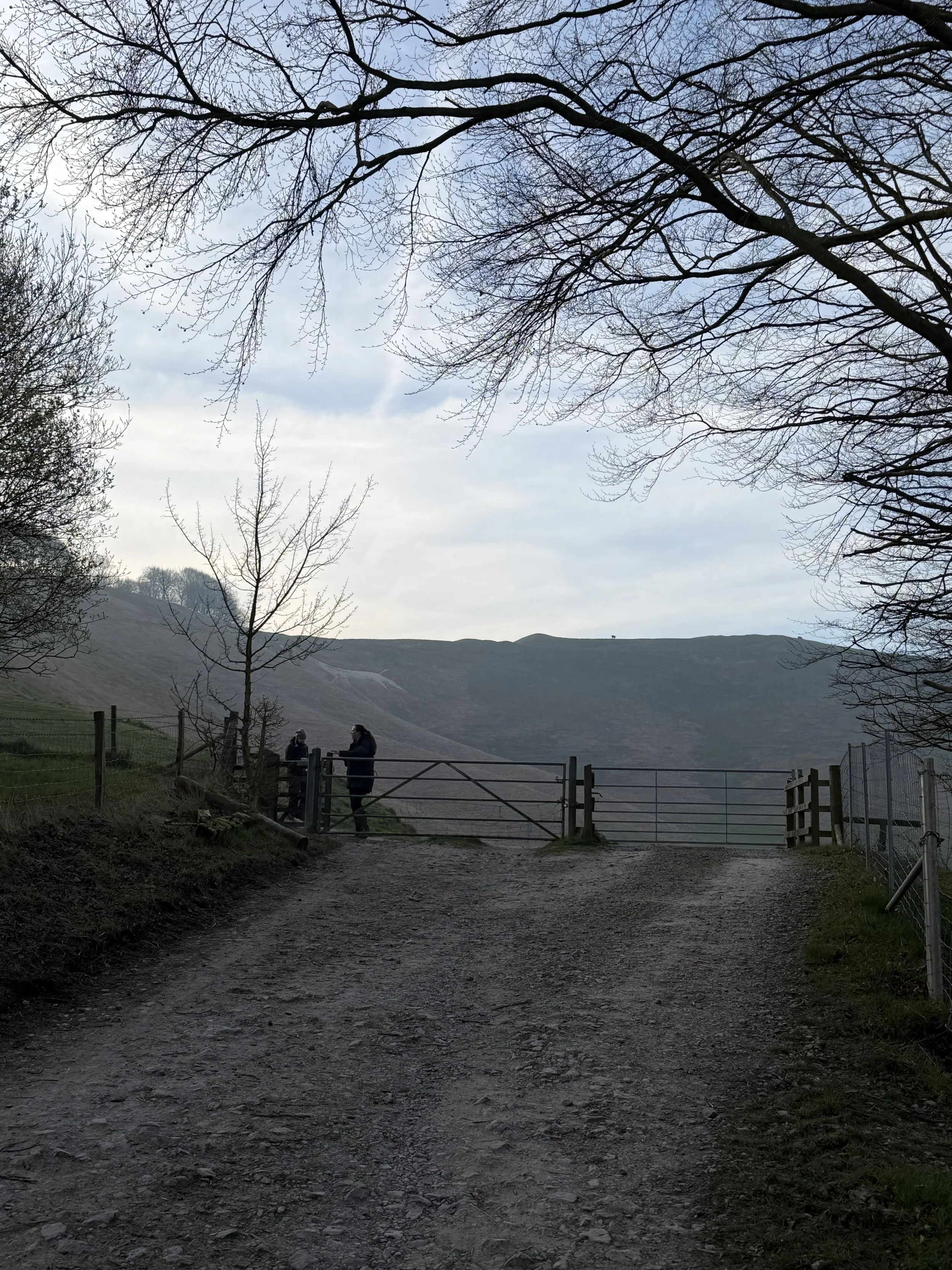 Two people standing by a gate on a dirt path in a rural hilly landscape with leafless trees and a cloudy sky.