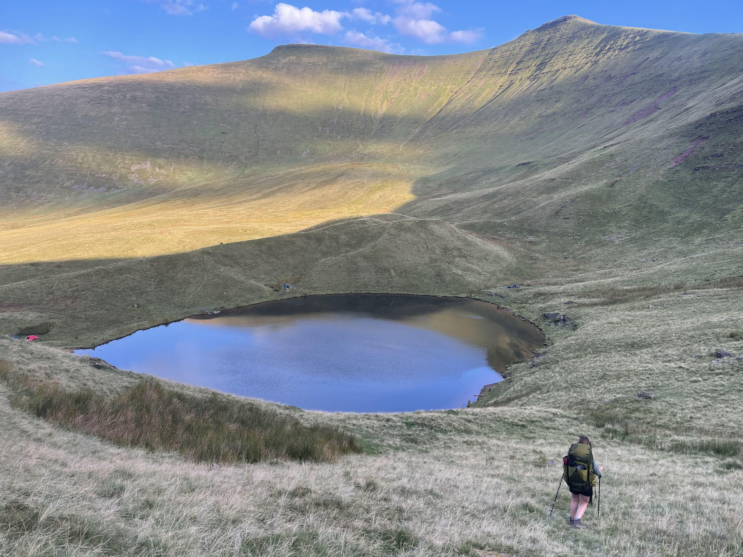 A hiker with a backpack and walking poles walking down a grassy hill towards a small lake in a mountainous landscape under a blue sky with some clouds.