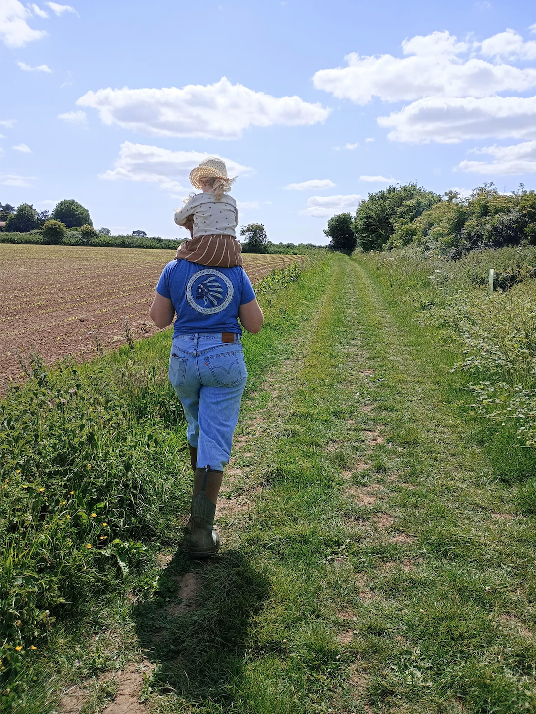 Person walking down a grassy rural path with a young girl sitting on their shoulders, clear blue sky with scattered clouds, green fields and trees on either side.