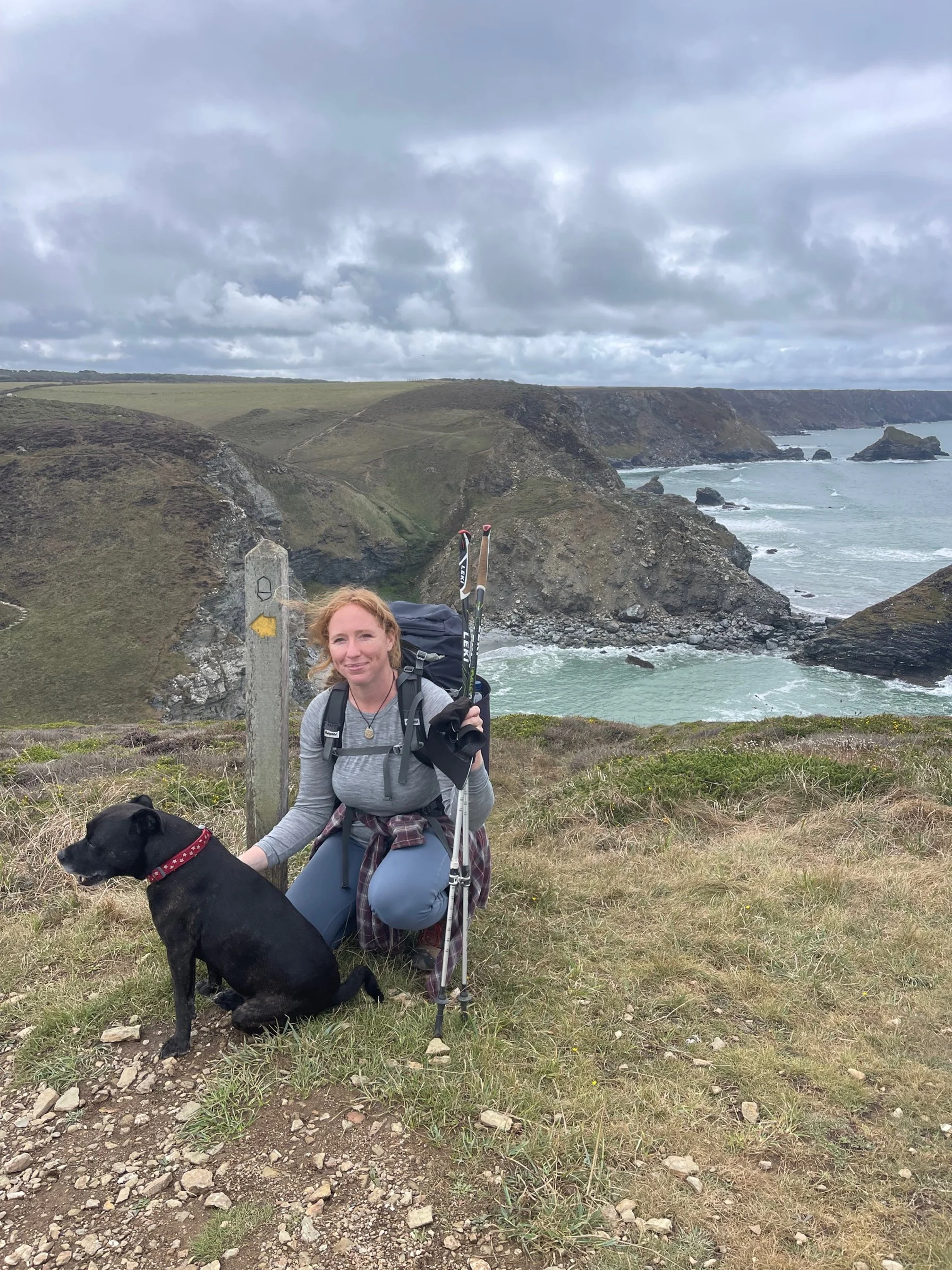 A woman with a backpack and hiking gear kneeling next to a black dog on a grassy cliff overlooking the ocean and rugged coastline under cloudy skies.