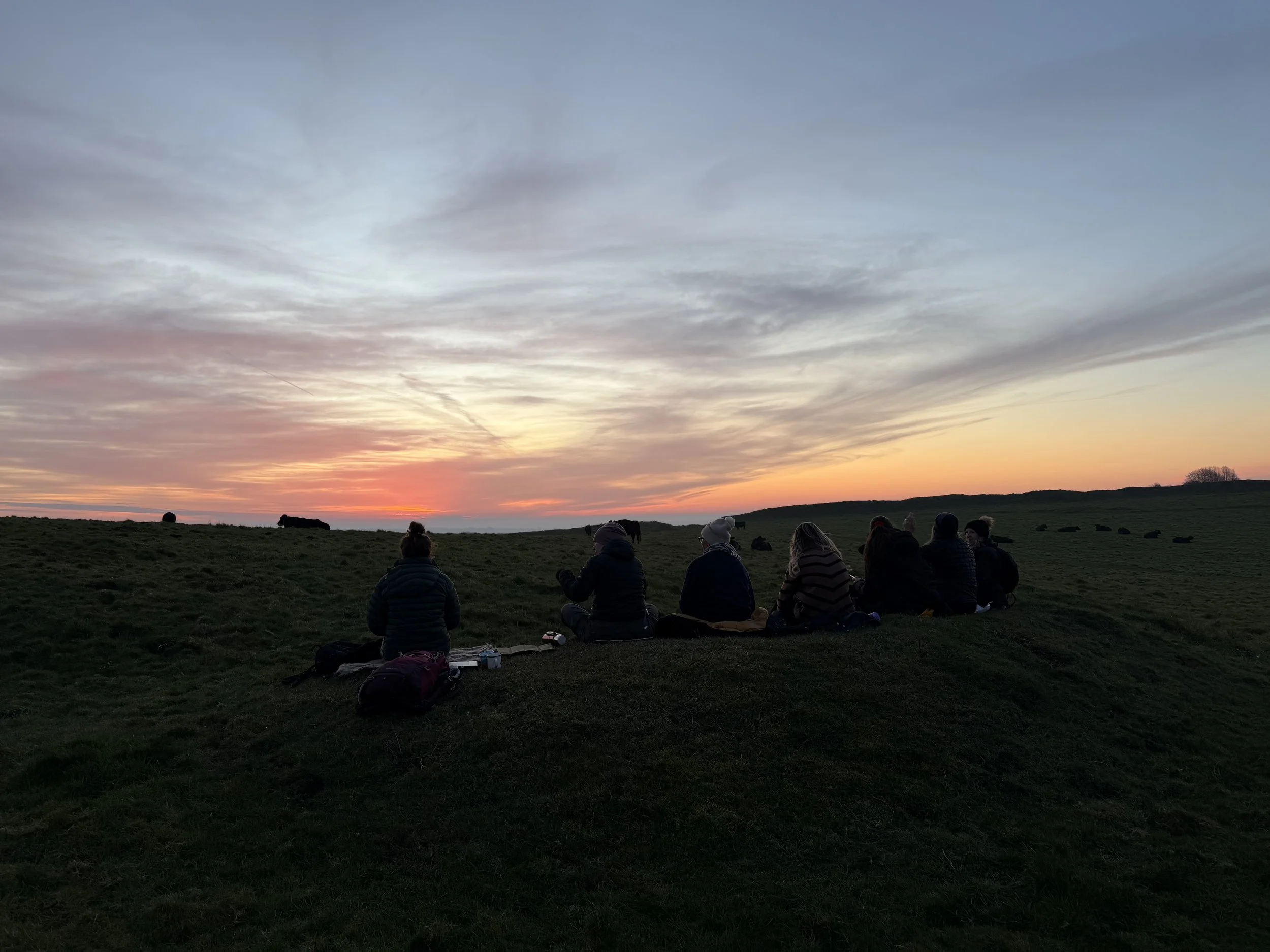 A group of people sitting on a grassy hill at sunset, watching the sky with clouds and a colorful horizon, some with backpacks and belongings beside them.