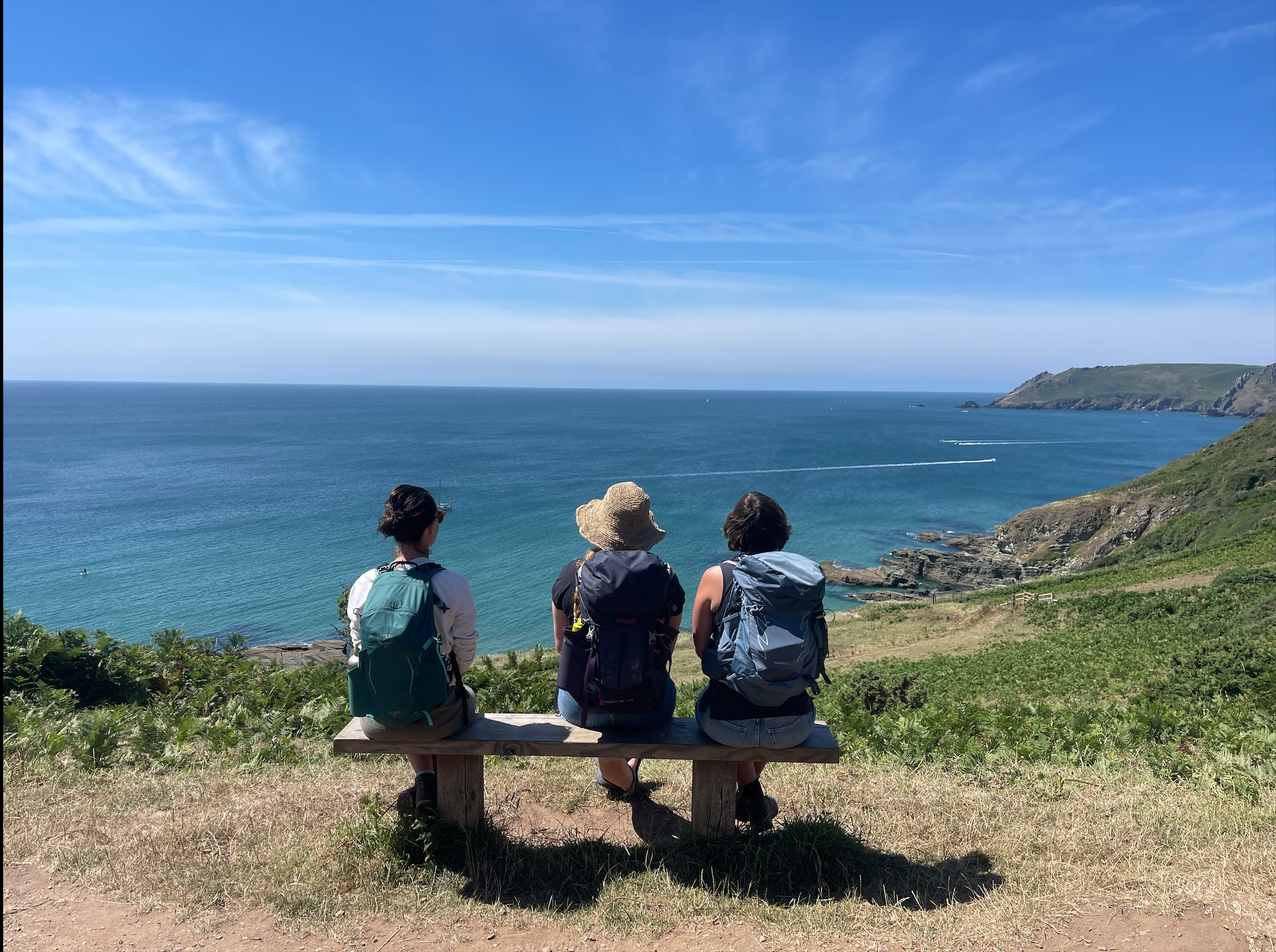 Three women with backpacks sitting on a bench overlooking a coastal landscape with water, cliffs, and a blue sky.