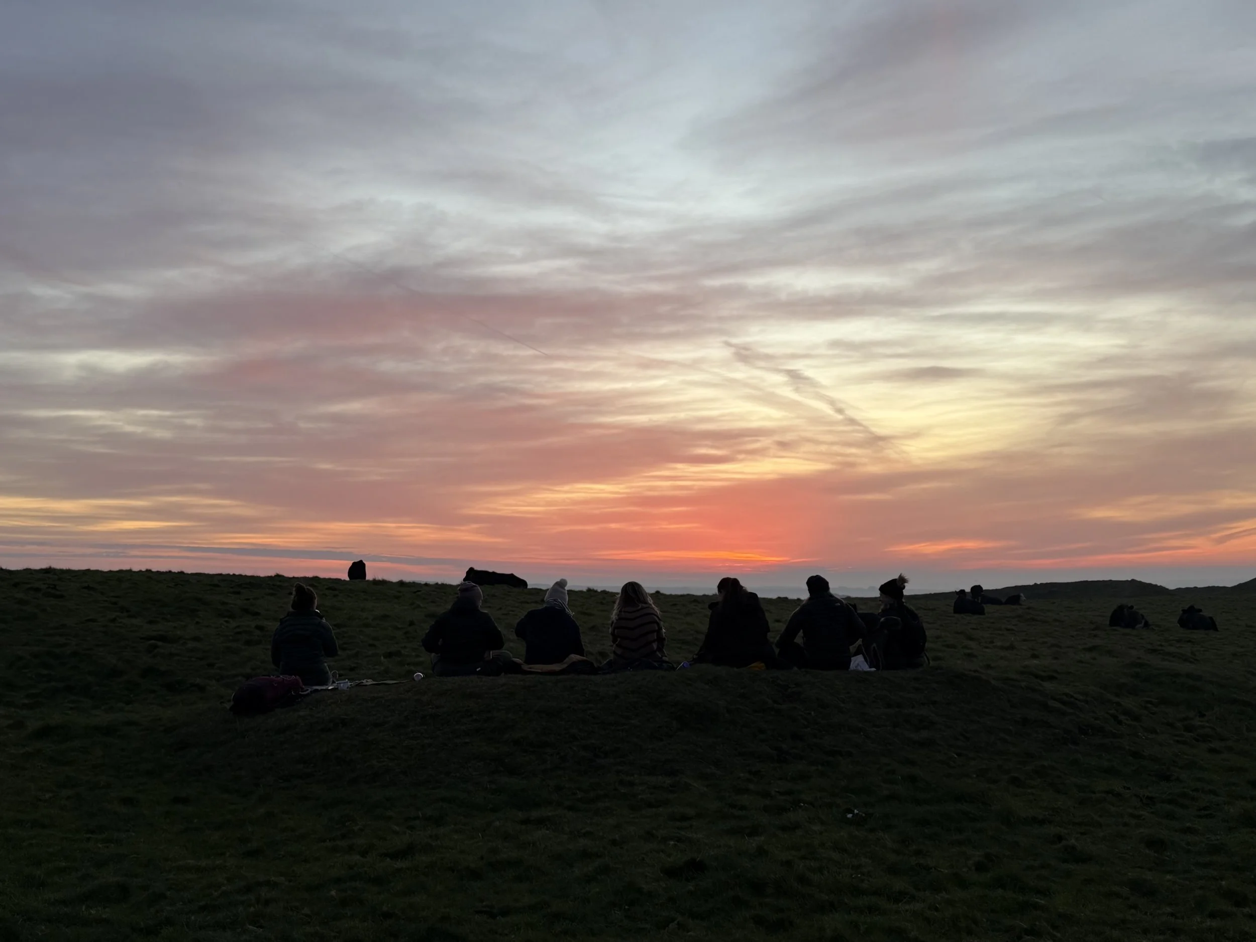 Group of people sitting on grass hill at sunset with colorful sky.