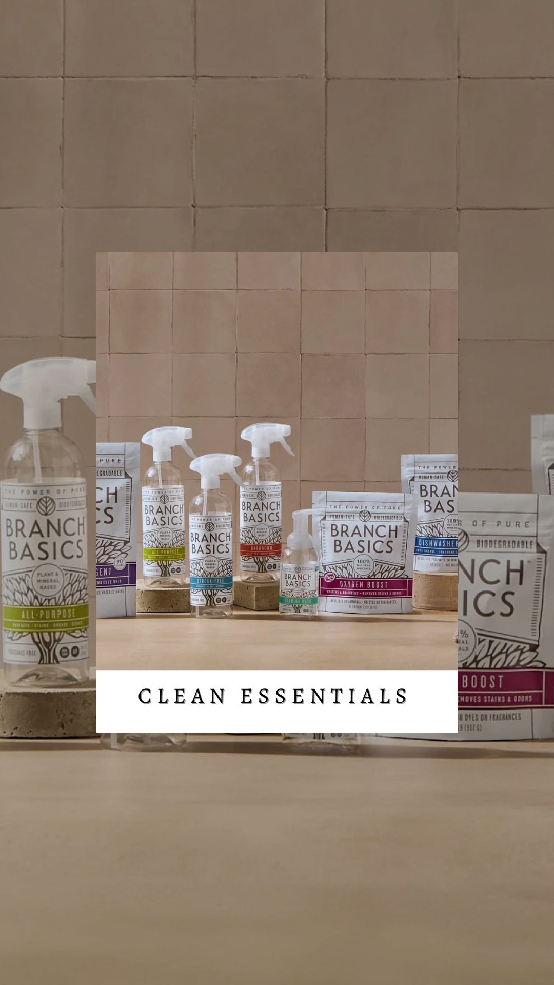 A collection of plant-based cleaning products from Branch Basics on a wooden surface with a beige tiled wall in the background. The products include spray bottles and refill pouches labeled for various cleaning purposes. The image has a white overlay with black text that reads 'CLEAN ESSENTIALS'.
