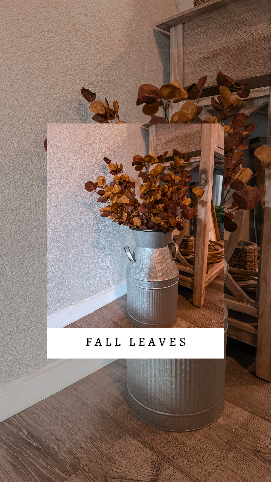 A rustic metal vase holding fall leaves with brown, yellow, and reddish hues, placed on a wooden floor next to a wooden shelf or cabinet.