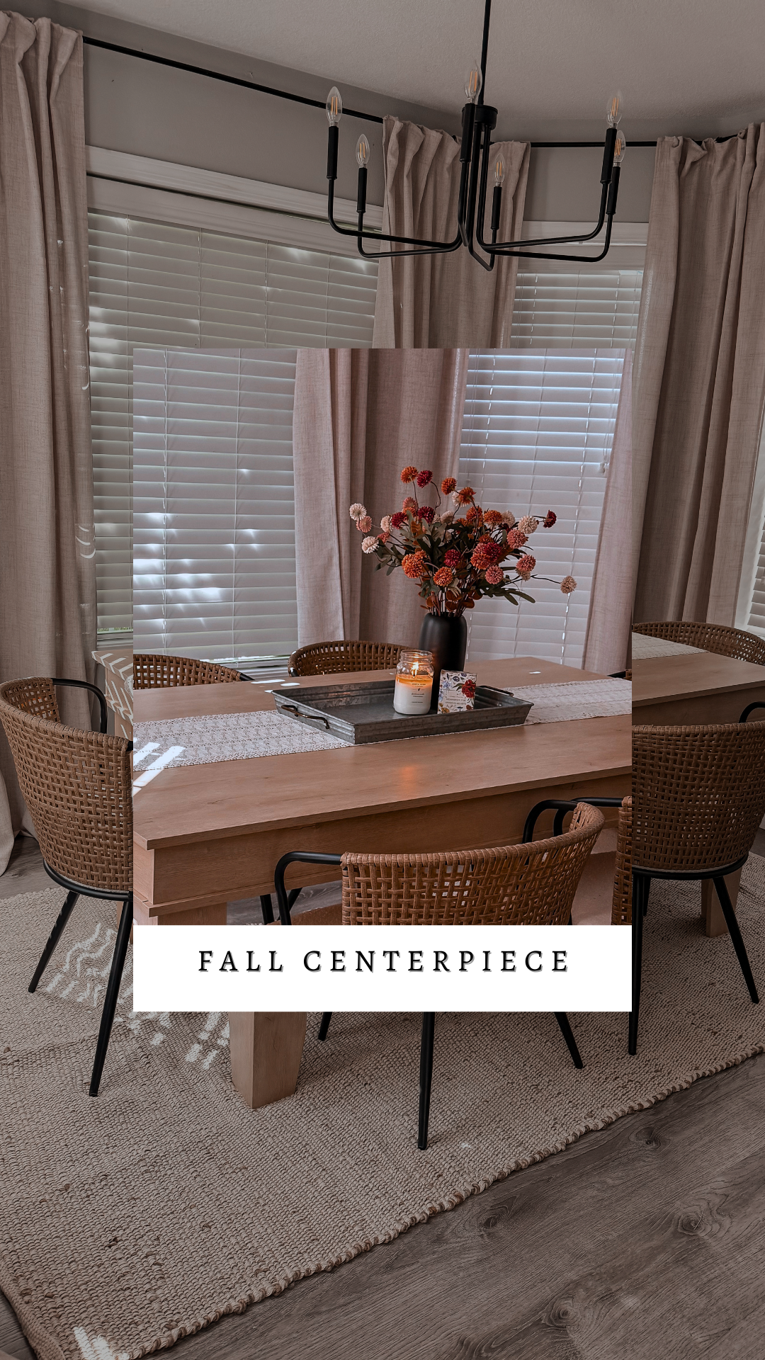 A dining room with a wooden table, woven chairs, a pottery vase with flowers, a candle, and a fall-themed card, all under a black chandelier and surrounded by windows with blinds and beige curtains.