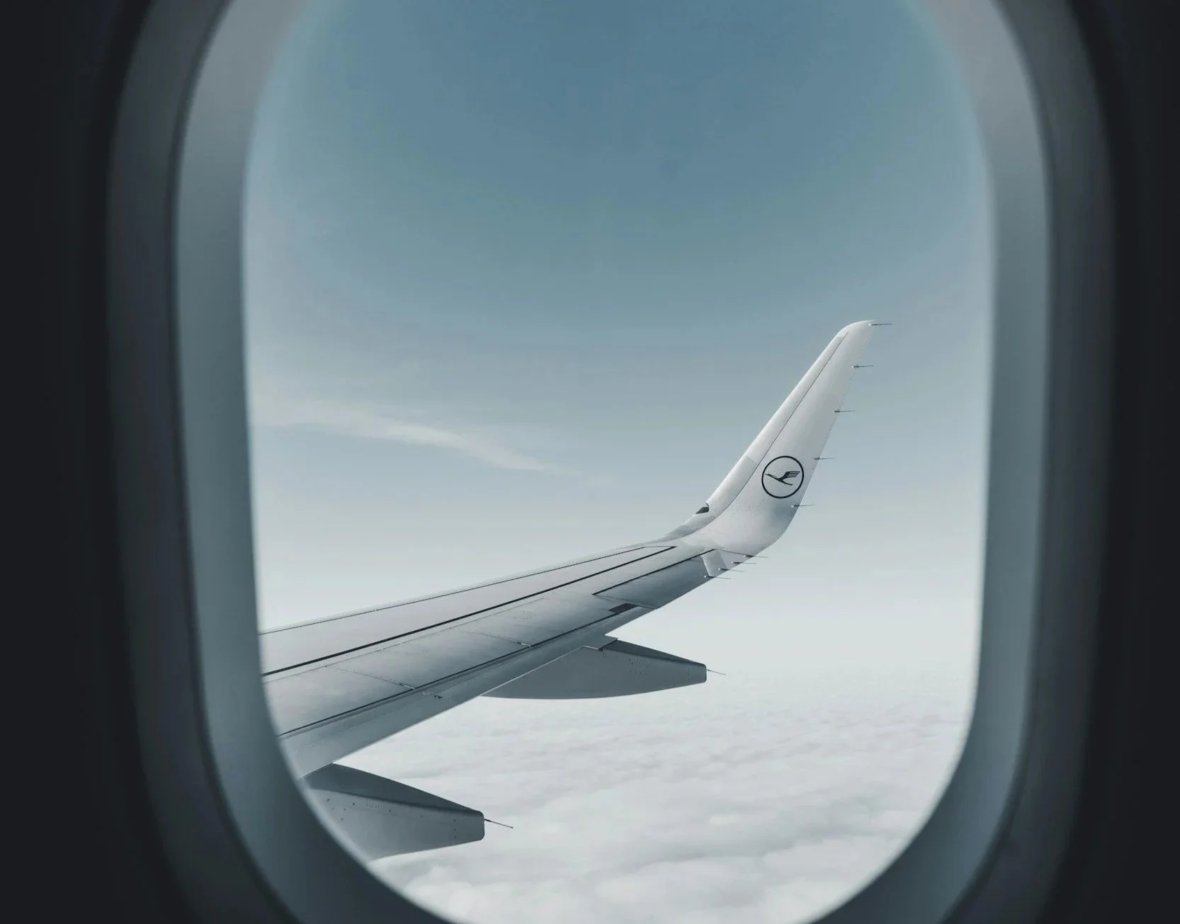 View of an airplane wing through an aircraft window, flying above the clouds on a clear day.