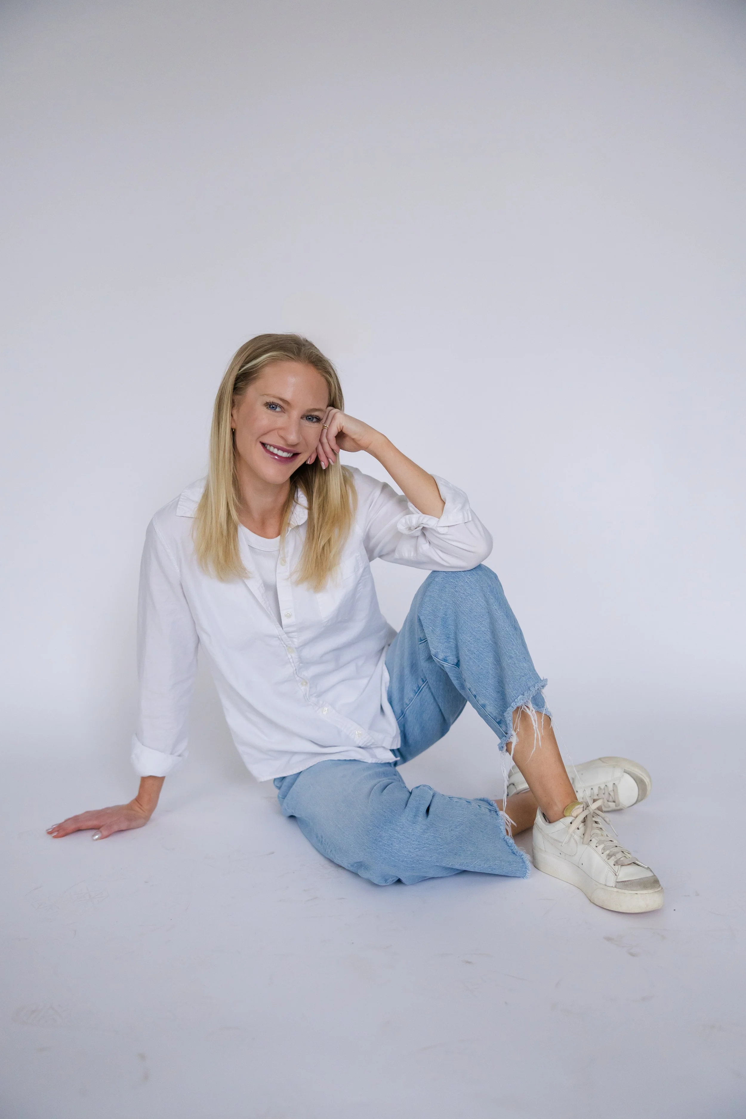 A young woman with blonde hair, wearing a white shirt, ripped jeans, and white sneakers, sitting on the floor against a plain white background, smiling and looking at the camera.