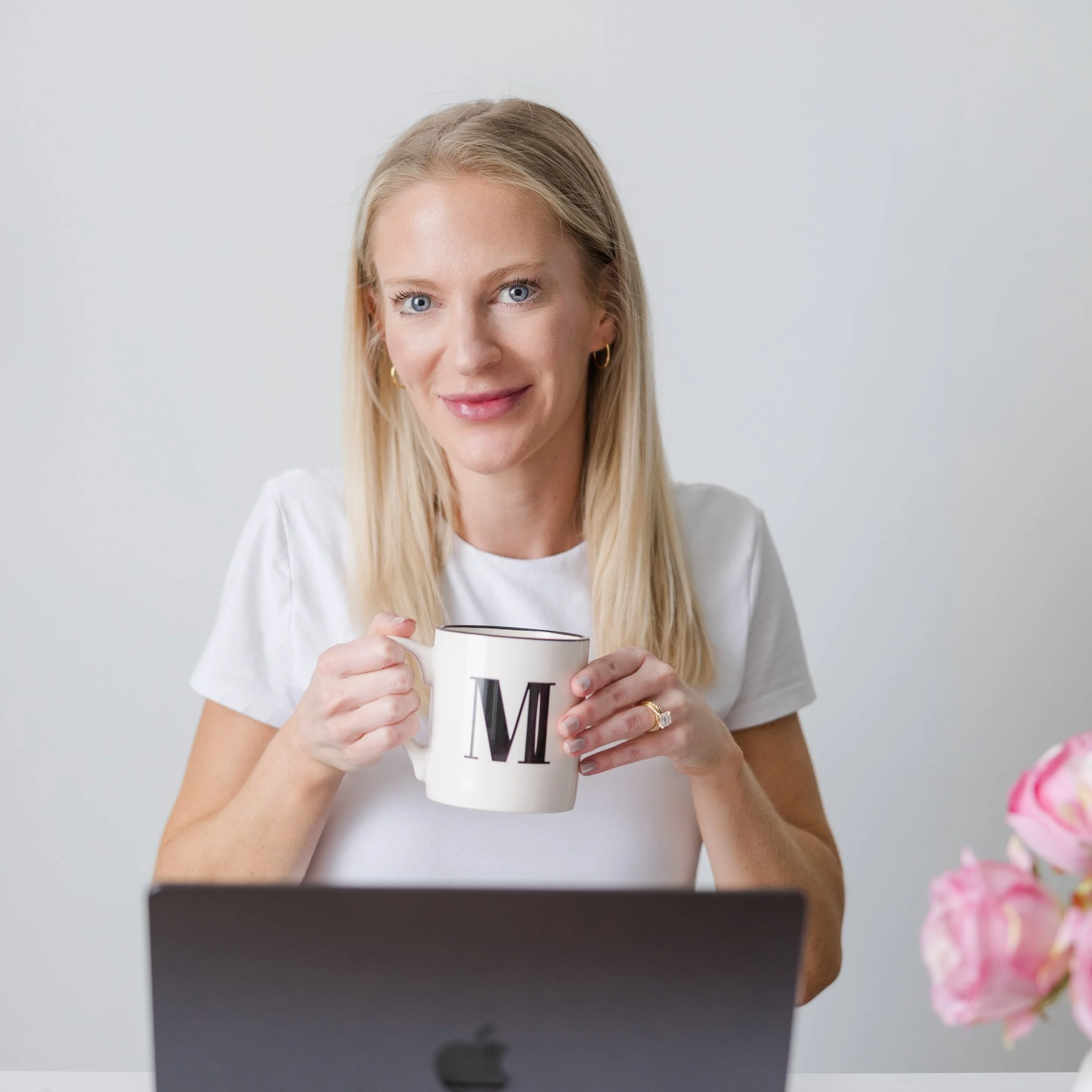 A woman with blonde hair and blue eyes smiling while holding a white mug with a black letter 'M' on it, sitting in front of a laptop with pink flowers on the right side.