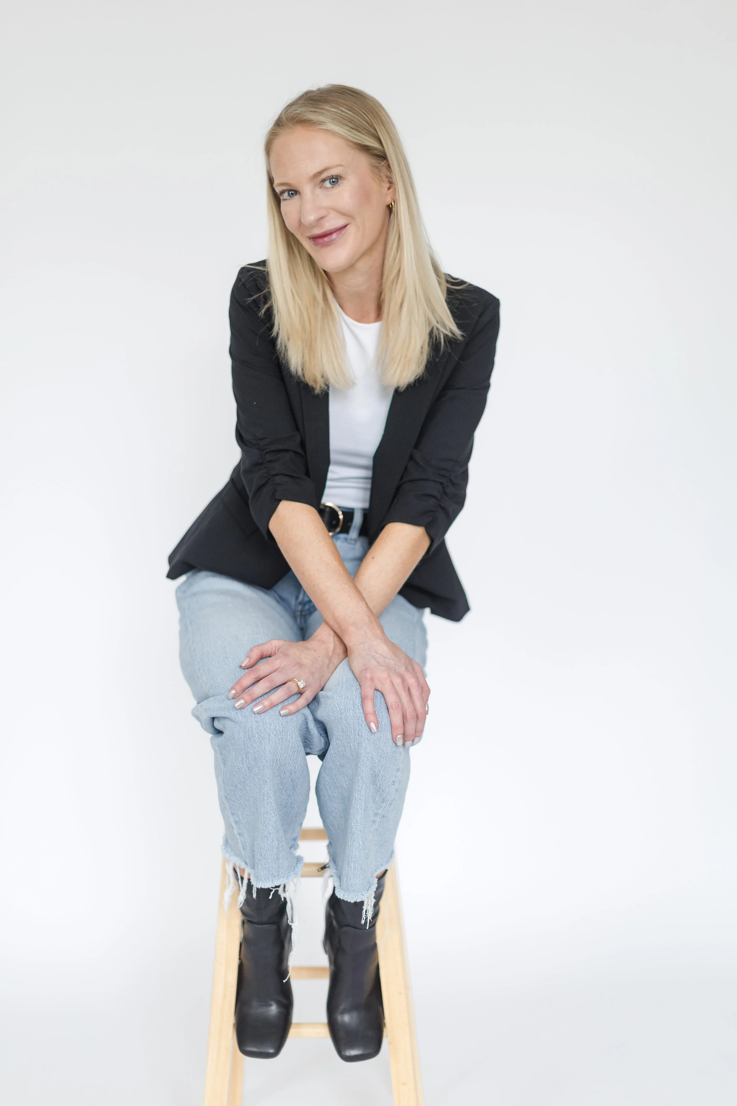 A woman with blonde hair sitting on a wooden stool, wearing a black blazer, white t-shirt, ripped jeans, and black boots, smiling at the camera against a plain white background.