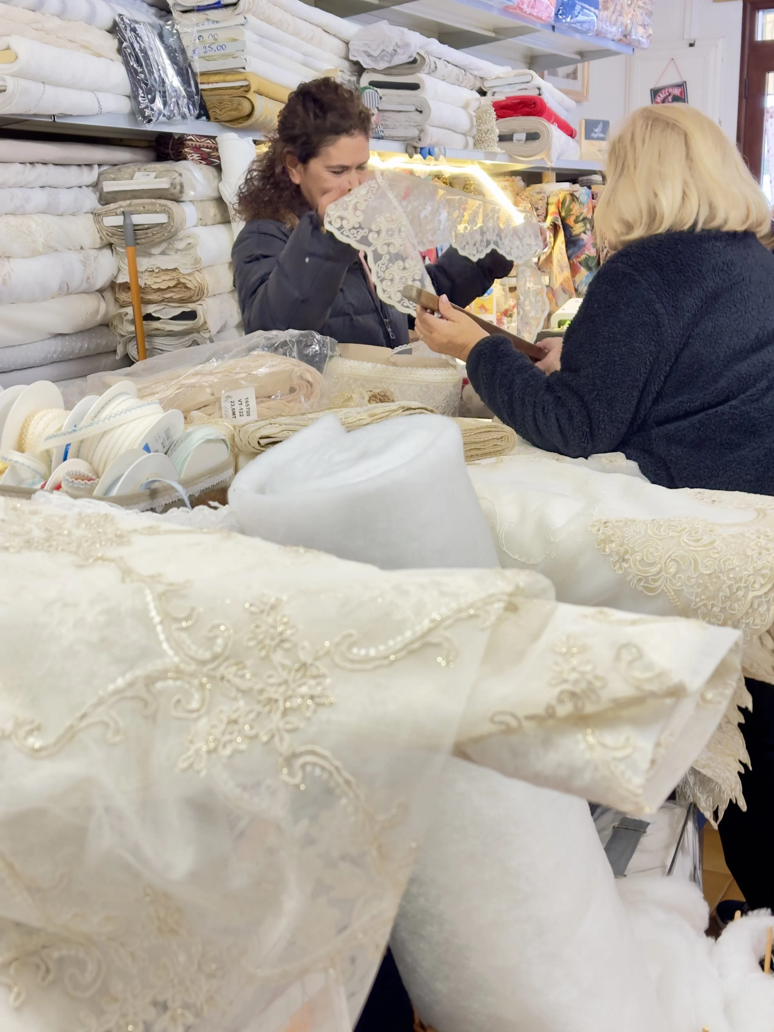 Due donne che si confrontano su tessuti e pizzi ricamati, con scaffali di tessuti colorati sullo sfondo.