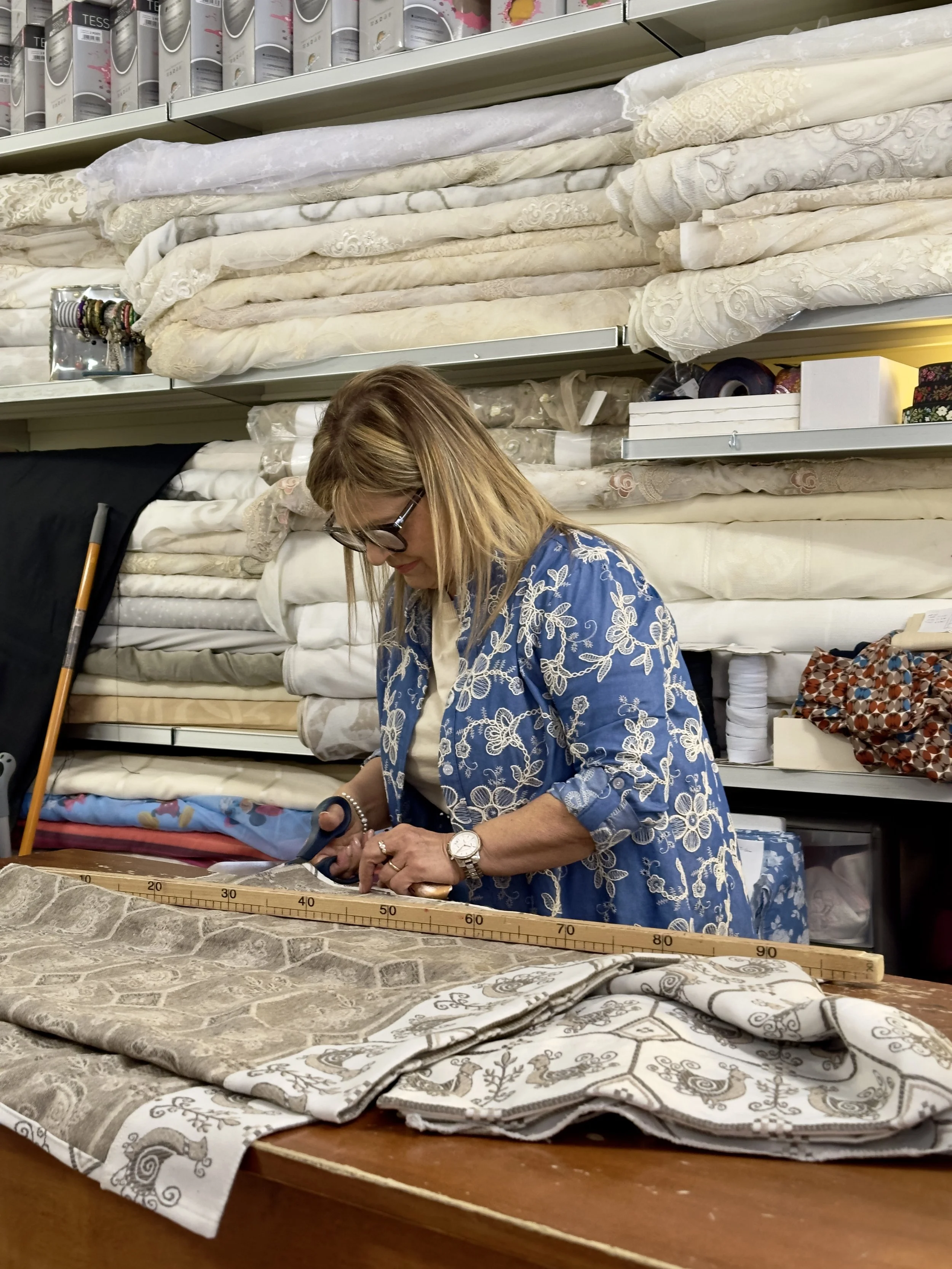 Gianna con occhiali e vestito blu con ricami bianche sta tagliando stoffa con delle forbici nel suo negozio di tessuti. Sul tavolo ci sono pezzi di tessuto e una squadra di misura in legno.