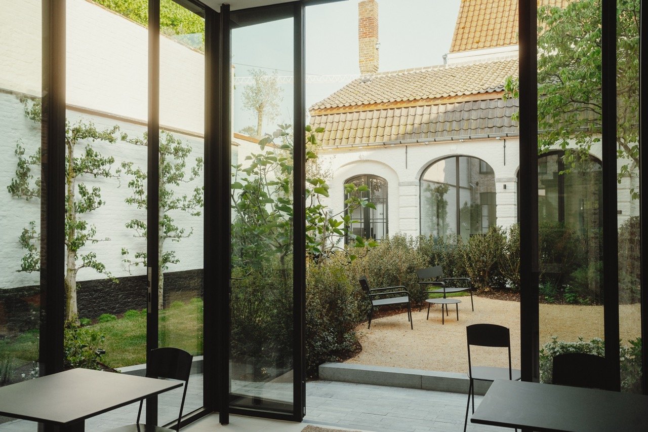 Binnenzicht naar een gezellig patio tuin met zitplaatsen, omgeven door een glazen muur, met een traditioneel wit huis op de achtergrond.