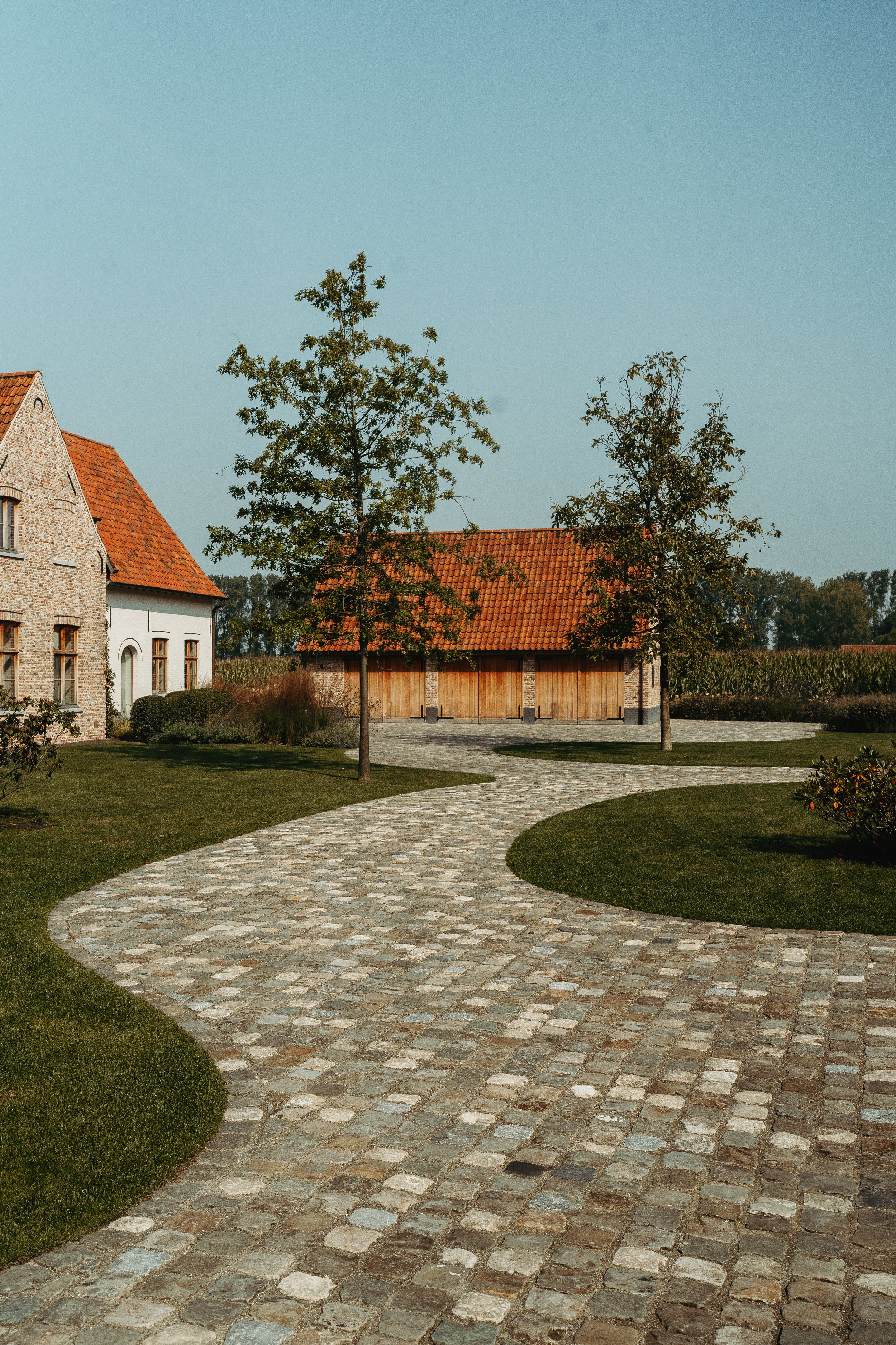 Een grindpad dat door een groene tuin slingert, met twee jonge bomen en een stenen gebouw met rood dak op de achtergrond.