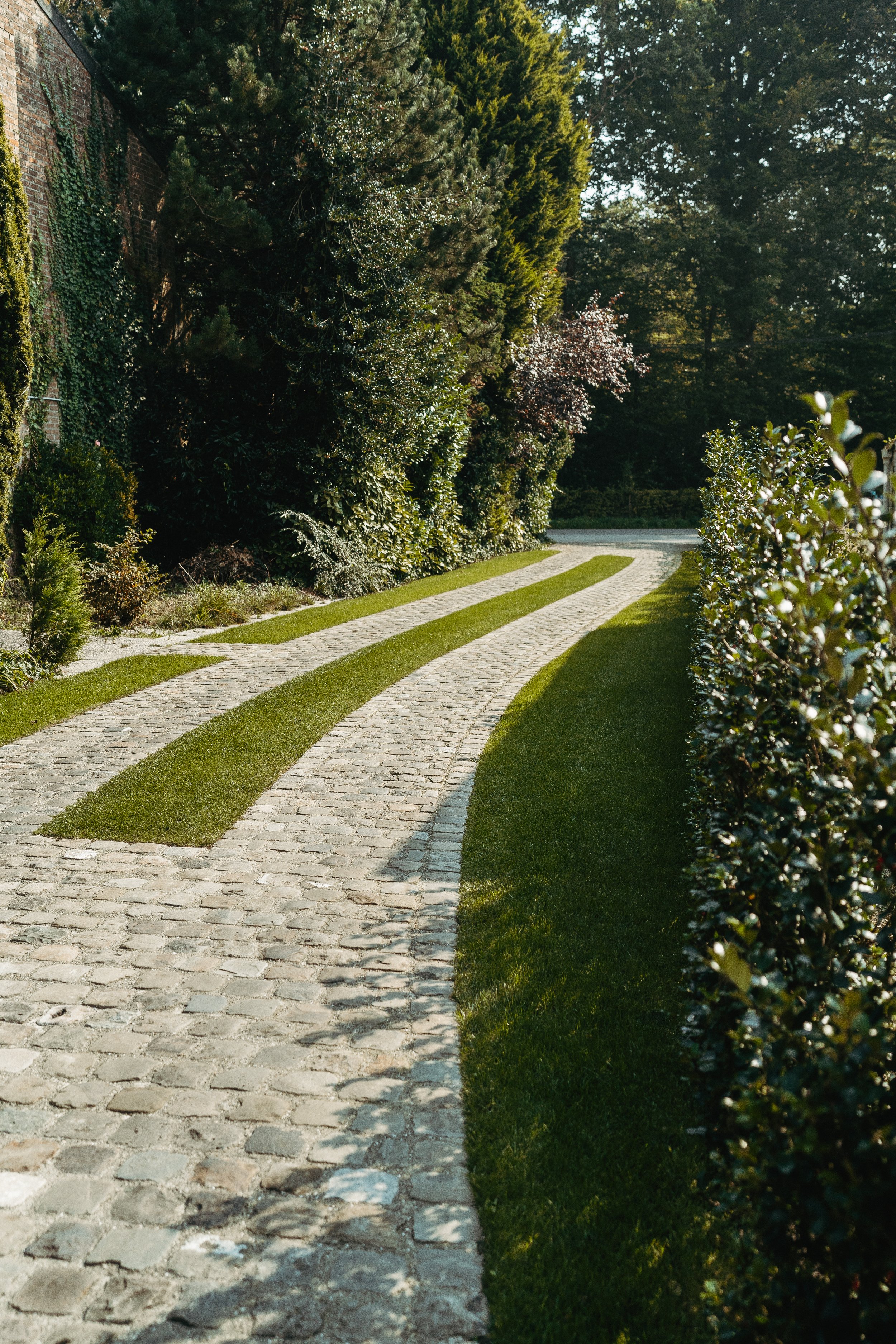 Een sierlijke kasseien oprit met decoratief gras tussen de stenen, omlind door groene struiken en bomen.
