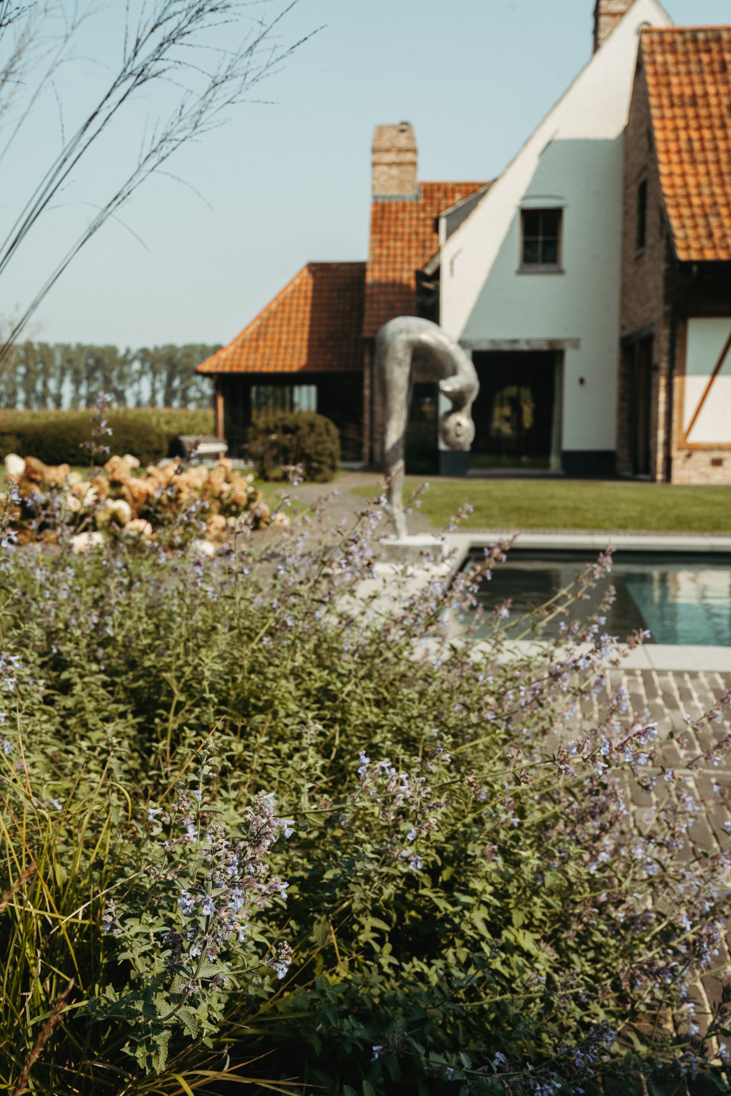 Tuin met bloemen, een zwembad, en een metalen beeld van een danseres voor een huis met een rood dak.