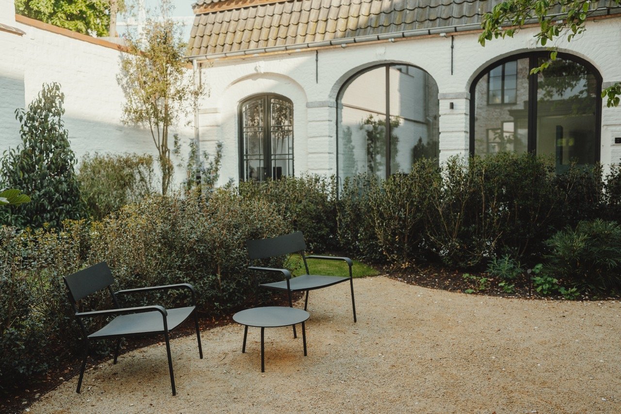 Twee zwarte tuinstoelen met een kleine ronde tafel op een grindpad in een tuin met groene struiken en een wit gebouw op de achtergrond.