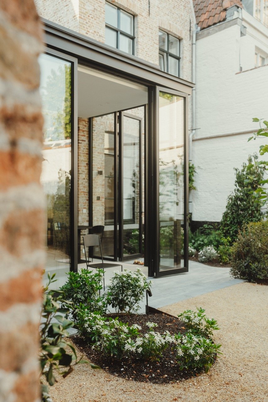 Moderne tuin met glazen terrasuitbreiding en bloembedden naast een witte bakstenen muur.