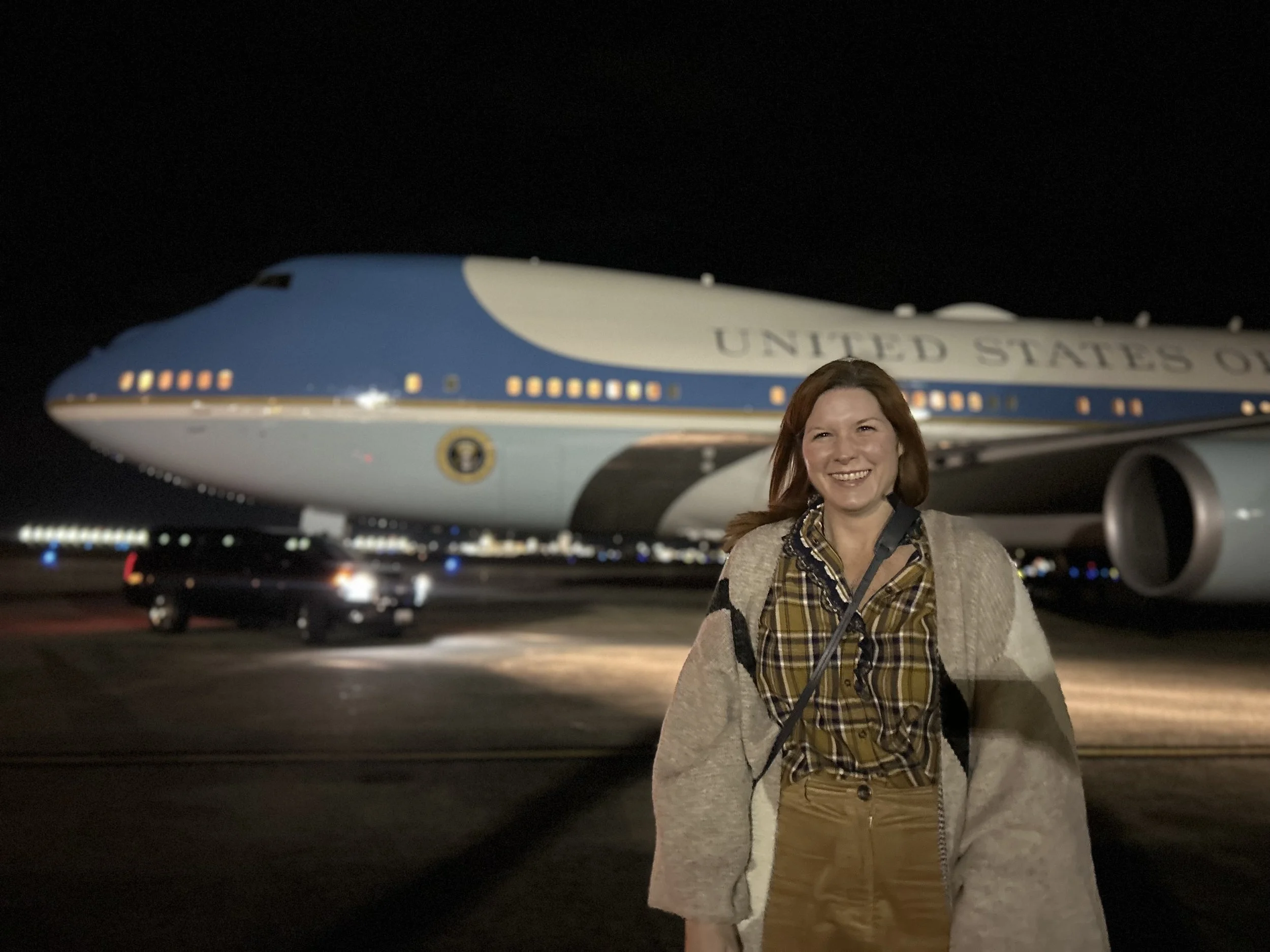 A woman smiling in front of Air Force One at night, with the airplane's nose and engine visible in the background.