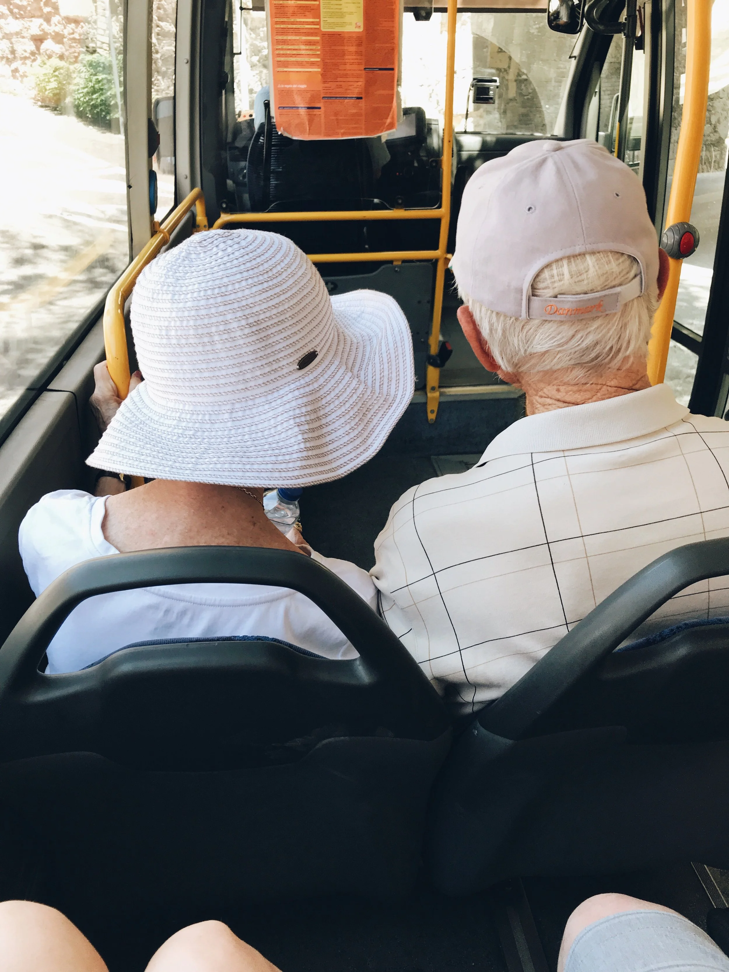 Two elderly people sitting on a bus, viewed from behind. The woman on the left is wearing a large white sun hat and a white top. The man on the right has white hair, a light-colored baseball cap, and a light-colored checkered shirt.