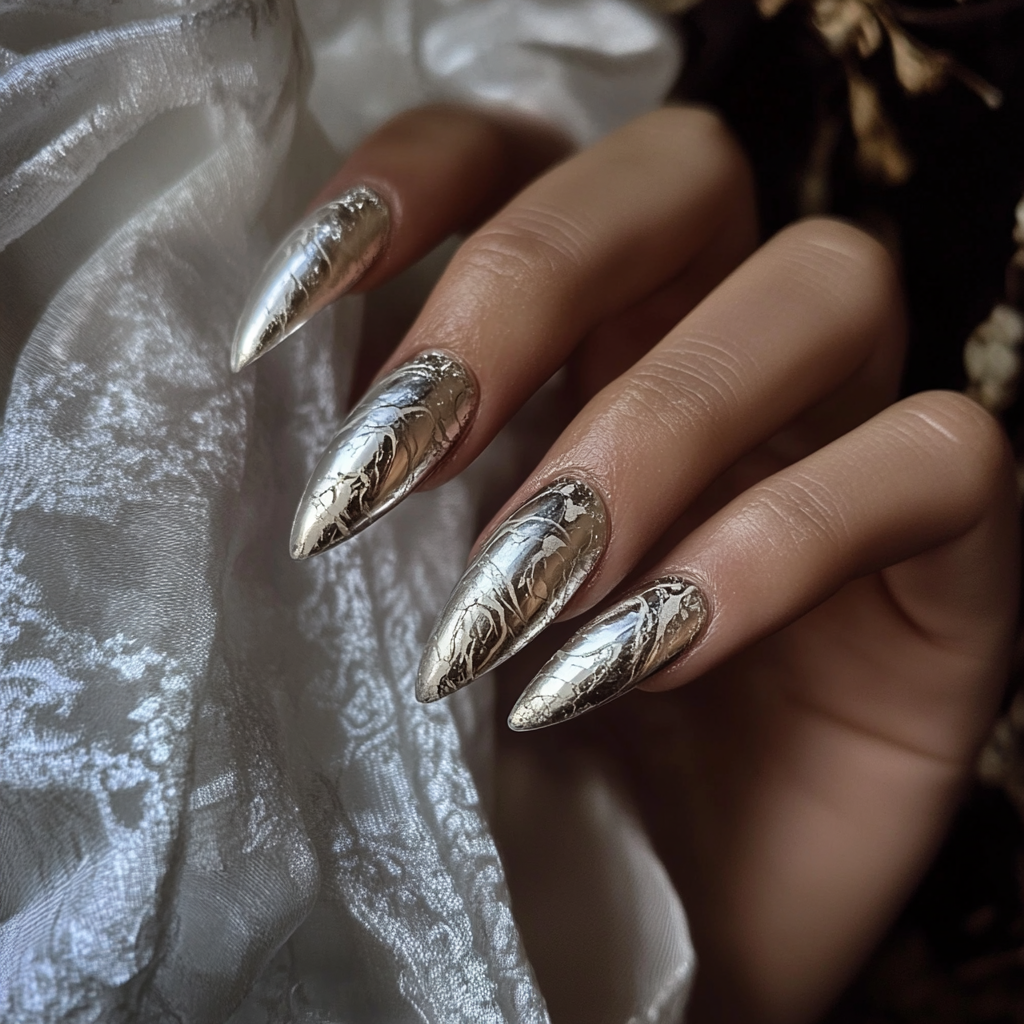 Close-up of a hand with long, pointed nails decorated with a metallic silver and black crackle design, resting on a silky, textured fabric.