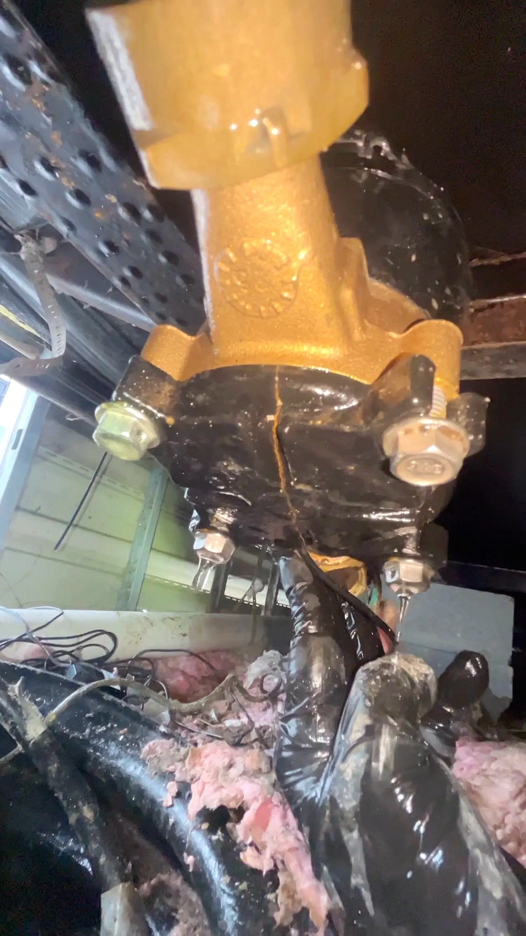 Close-up of a mobile home water meter leaking  because of cracked freeze plate with visible bolts and black and yellow coloring. Surrounding area shows pink insulation and wiring.