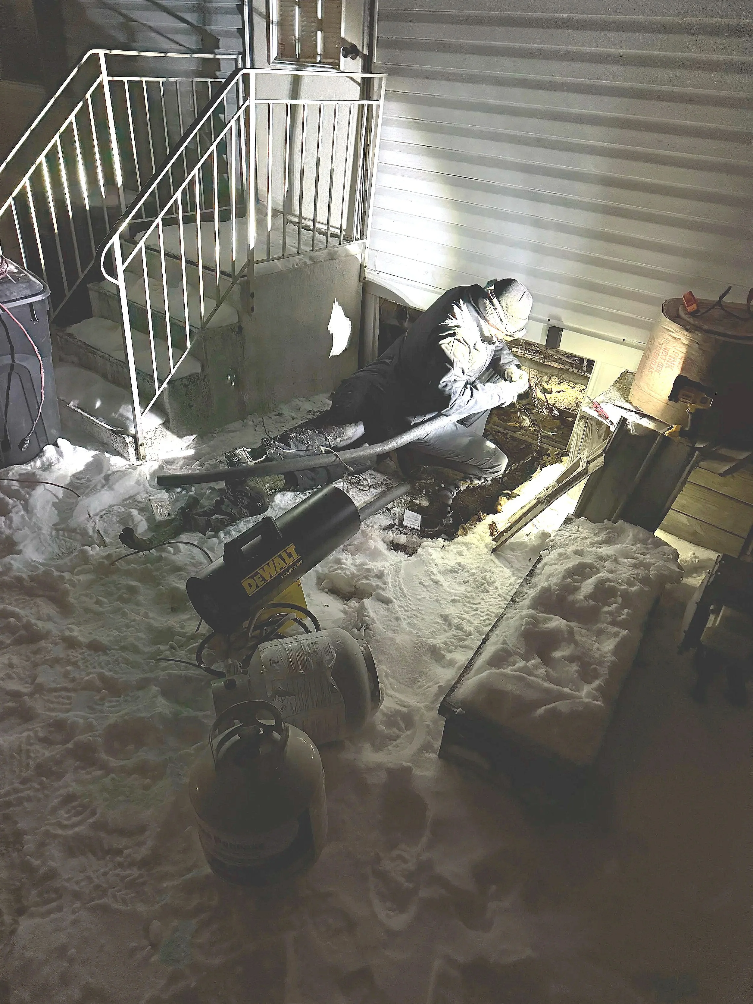 A worker using proper equipment to repair a frozen pipe in a crawlspace filled with snow, with a propane tank, and construction supplies nearby.