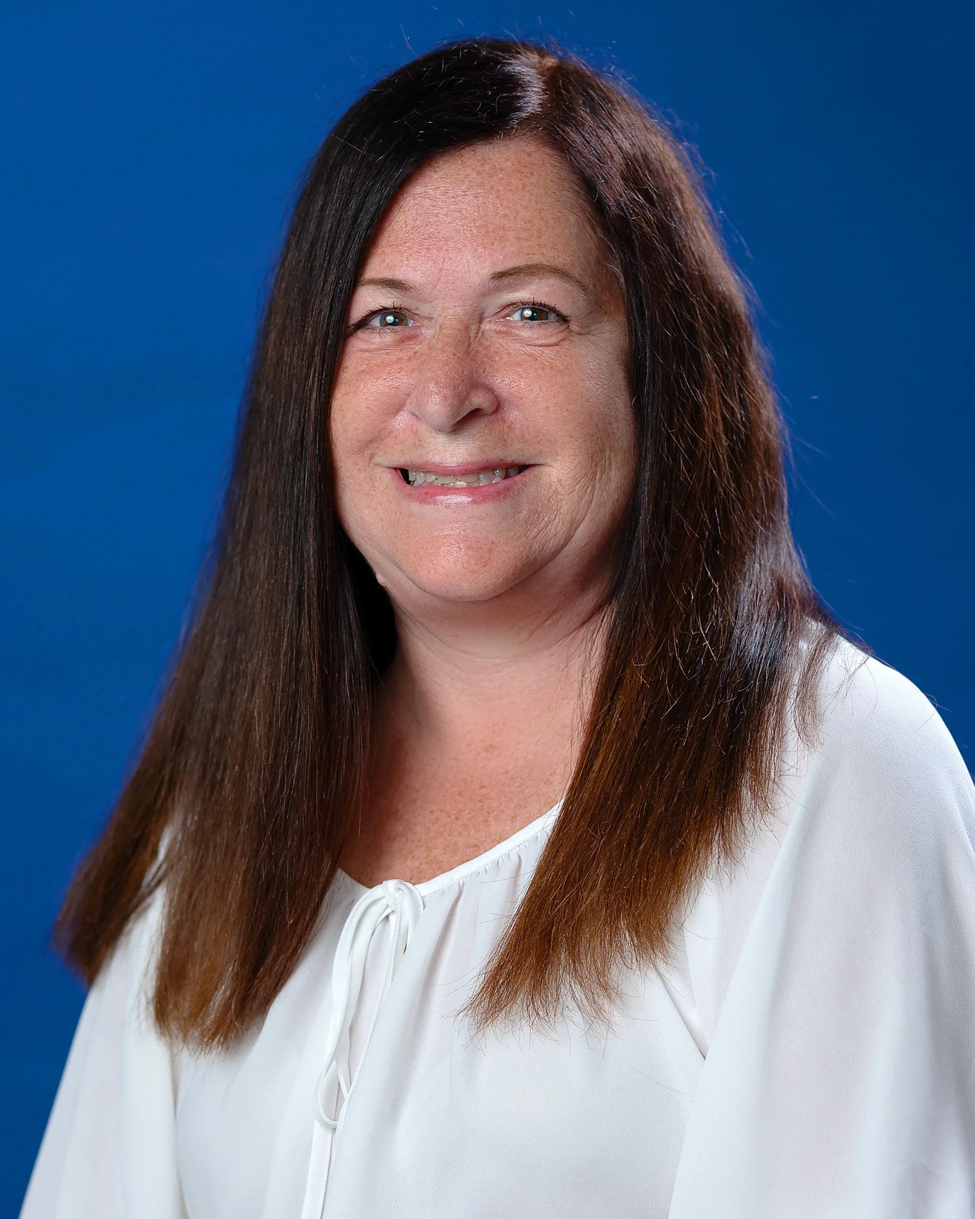 Woman smiling in a blue shirt with greenery in the background.