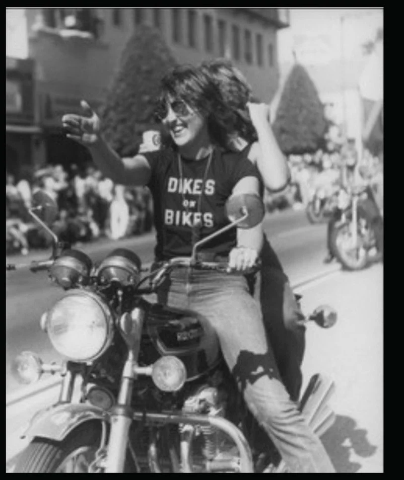 Woman with curly hair, wearing sunglasses and a t-shirt that says 'DIKES on BIKES', riding a motorcycle during a parade or event with onlookers in the background.