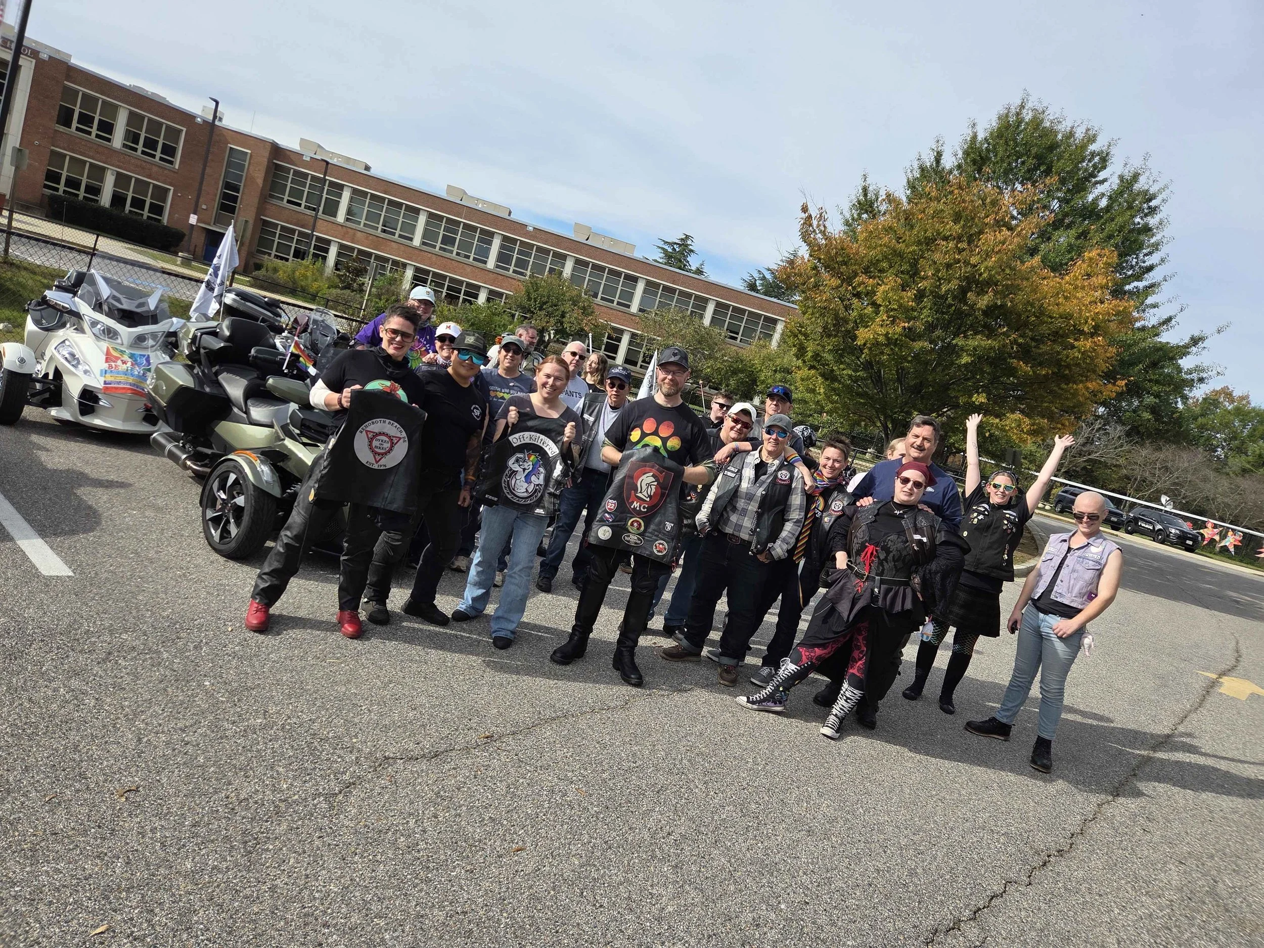 A group of people gathered outdoors near a school building, some wearing Pride flags and patches, standing next to three Yamaha motorcycles, with trees displaying fall foliage in the background.