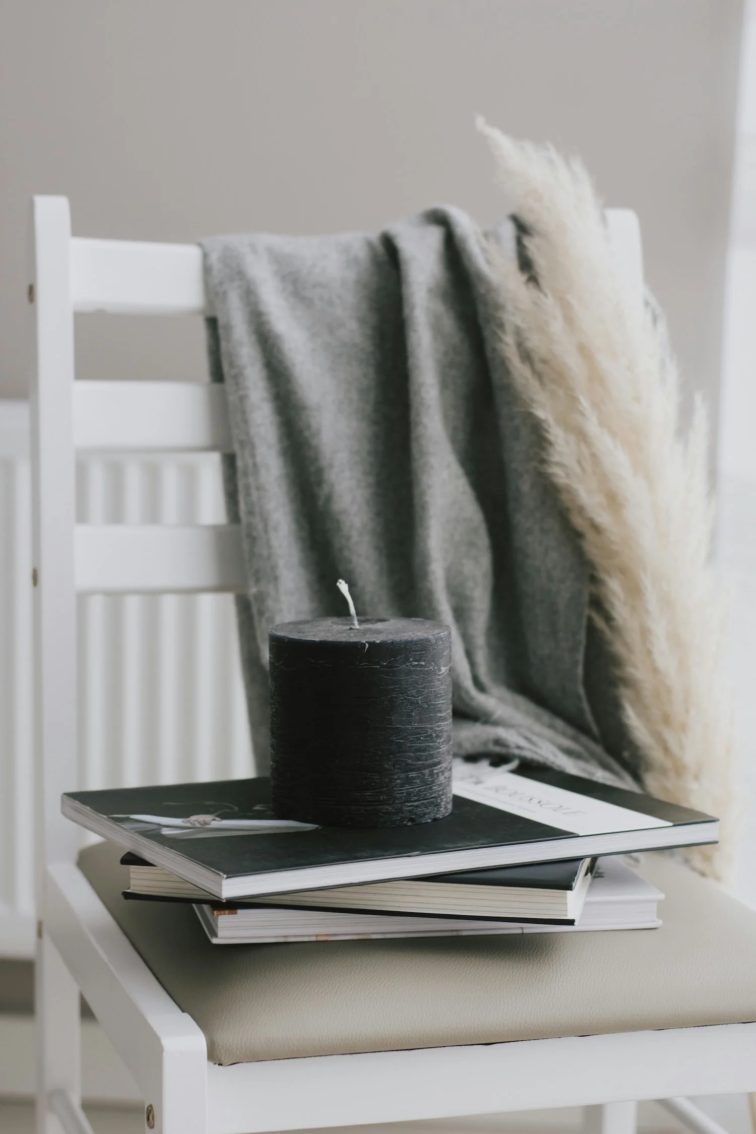 A white chair with a beige cushion, holding a stack of magazines, a black candle, and a gray and cream blanket draped over the backrest.