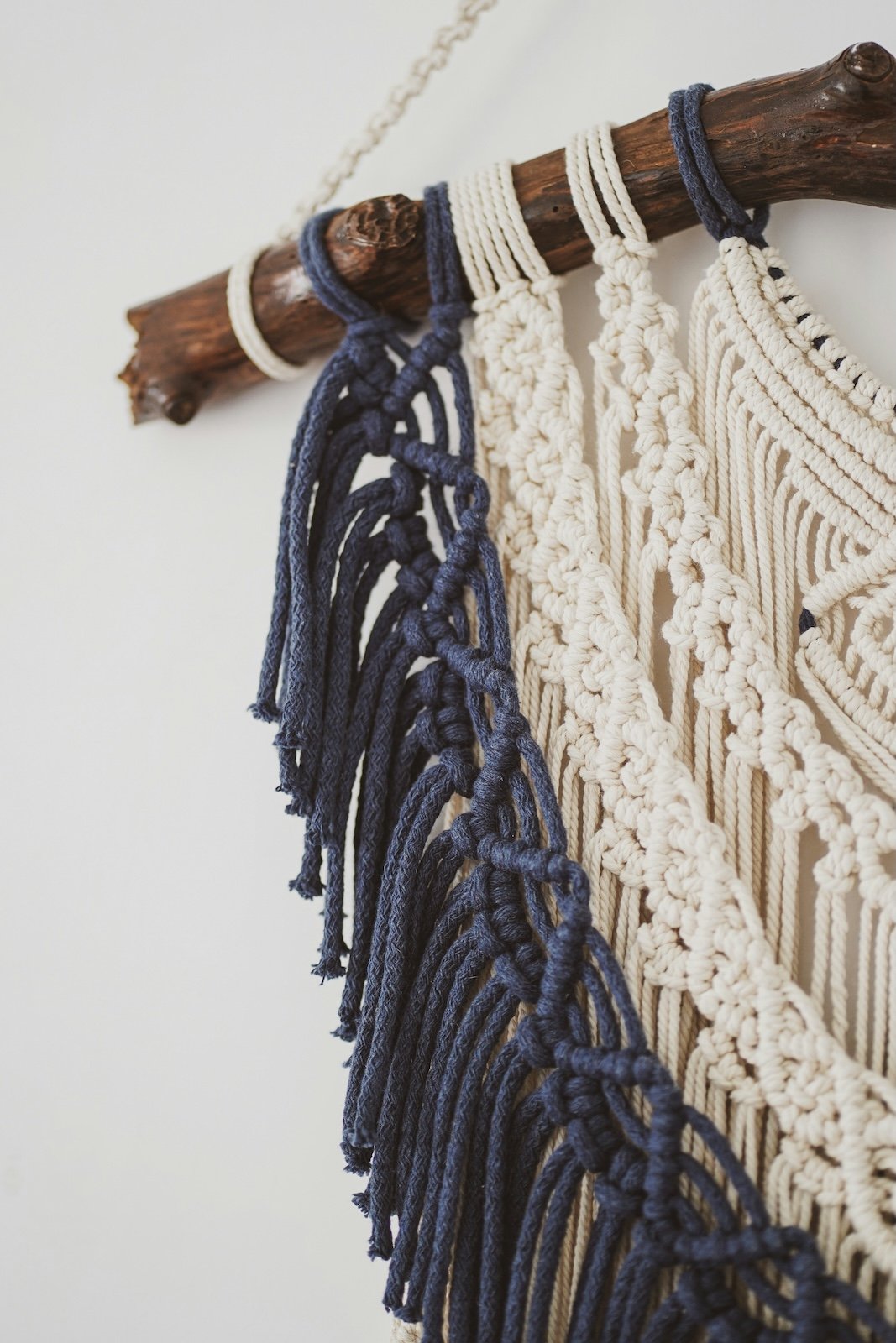 Close-up of a wall hanging with cream and navy blue knotted macrame designs secured on a wooden branch.