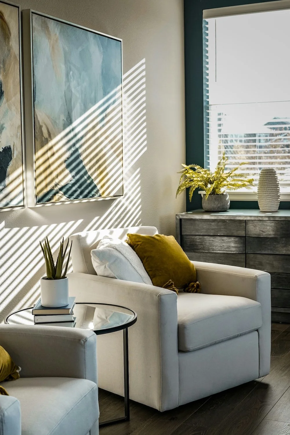 Living room with sunlight casting striped shadows on the wall, featuring a white armchair with a mustard yellow pillow, a small side table with a plant and books, a gray chest of drawers with plants and vases on top, and wall art.