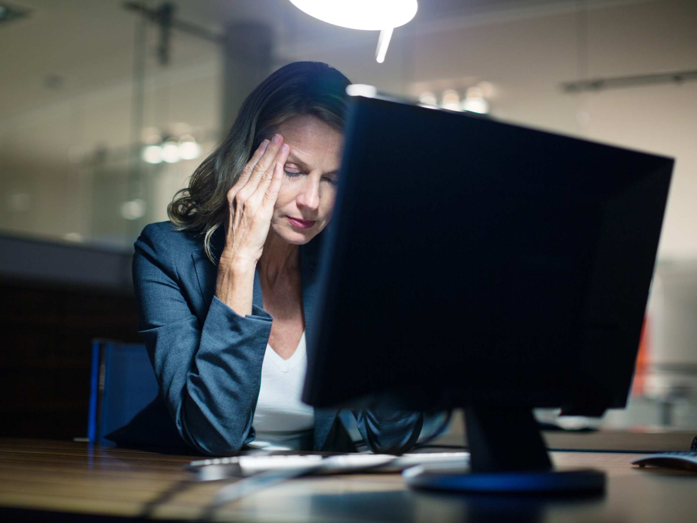 A woman in business attire sitting at a desk appears stressed, holding her head with her hand while looking at a computer monitor.